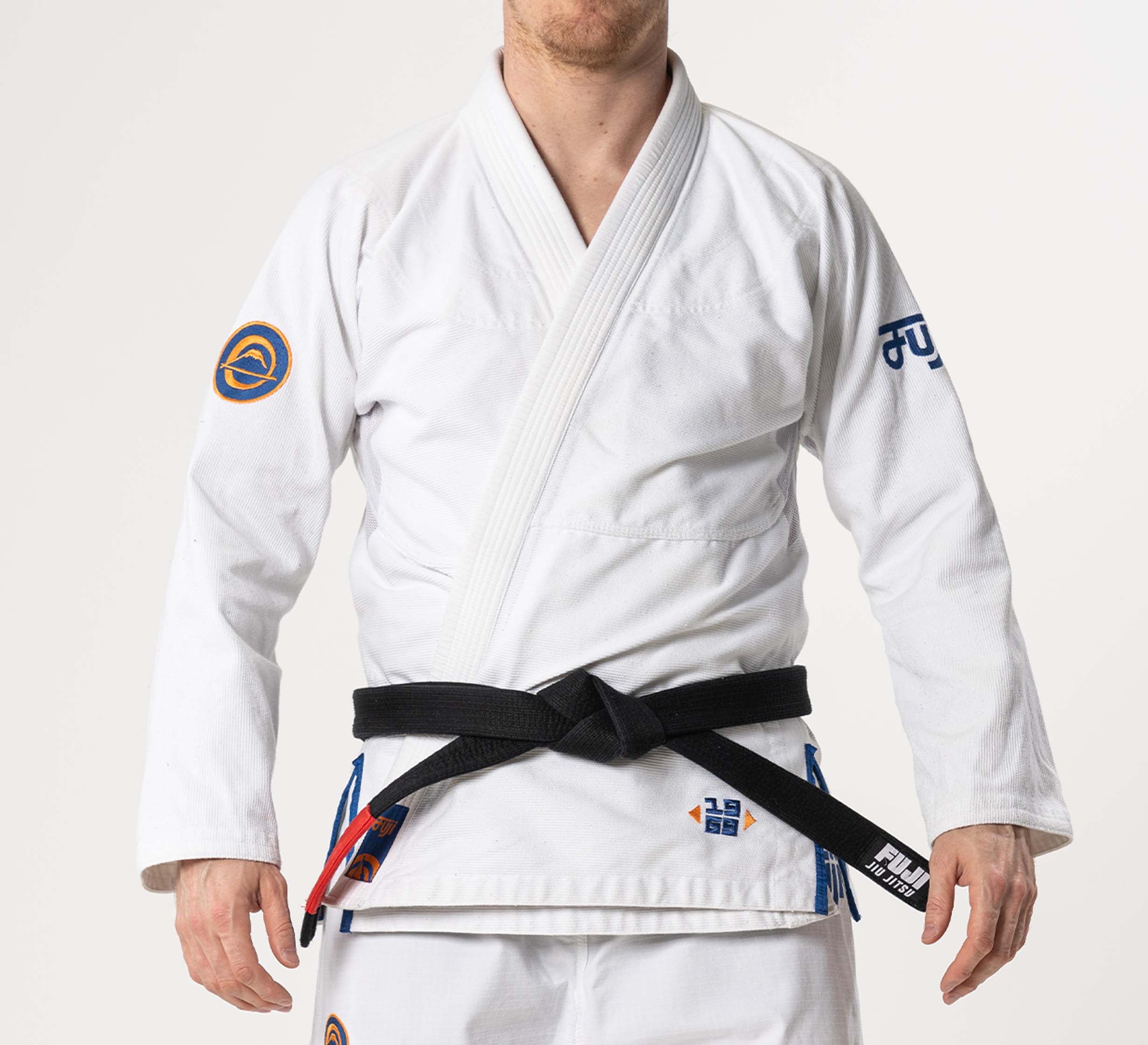 A person wearing the Flow-Tech BJJ Gi White/Navy/Orange with patches and a black belt stands against a plain background, head partially out of frame.