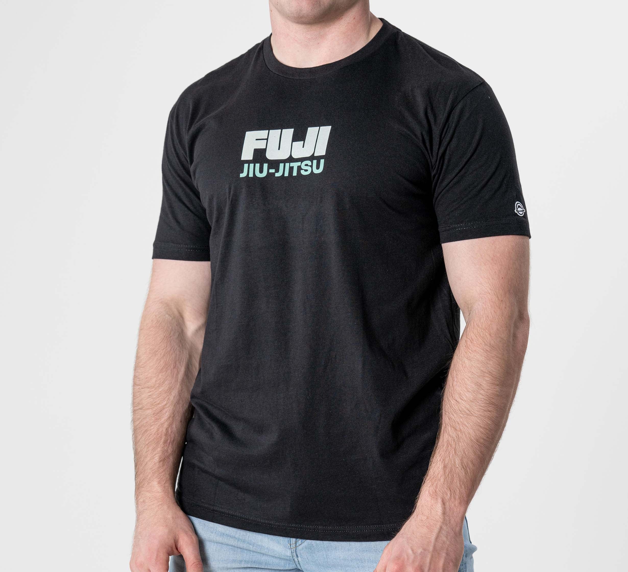 A man wearing the Black Belt Mentality T-Shirt Black, featuring FUJI JIU-JITSU in white and light blue lettering on the chest, stands against a plain light background.