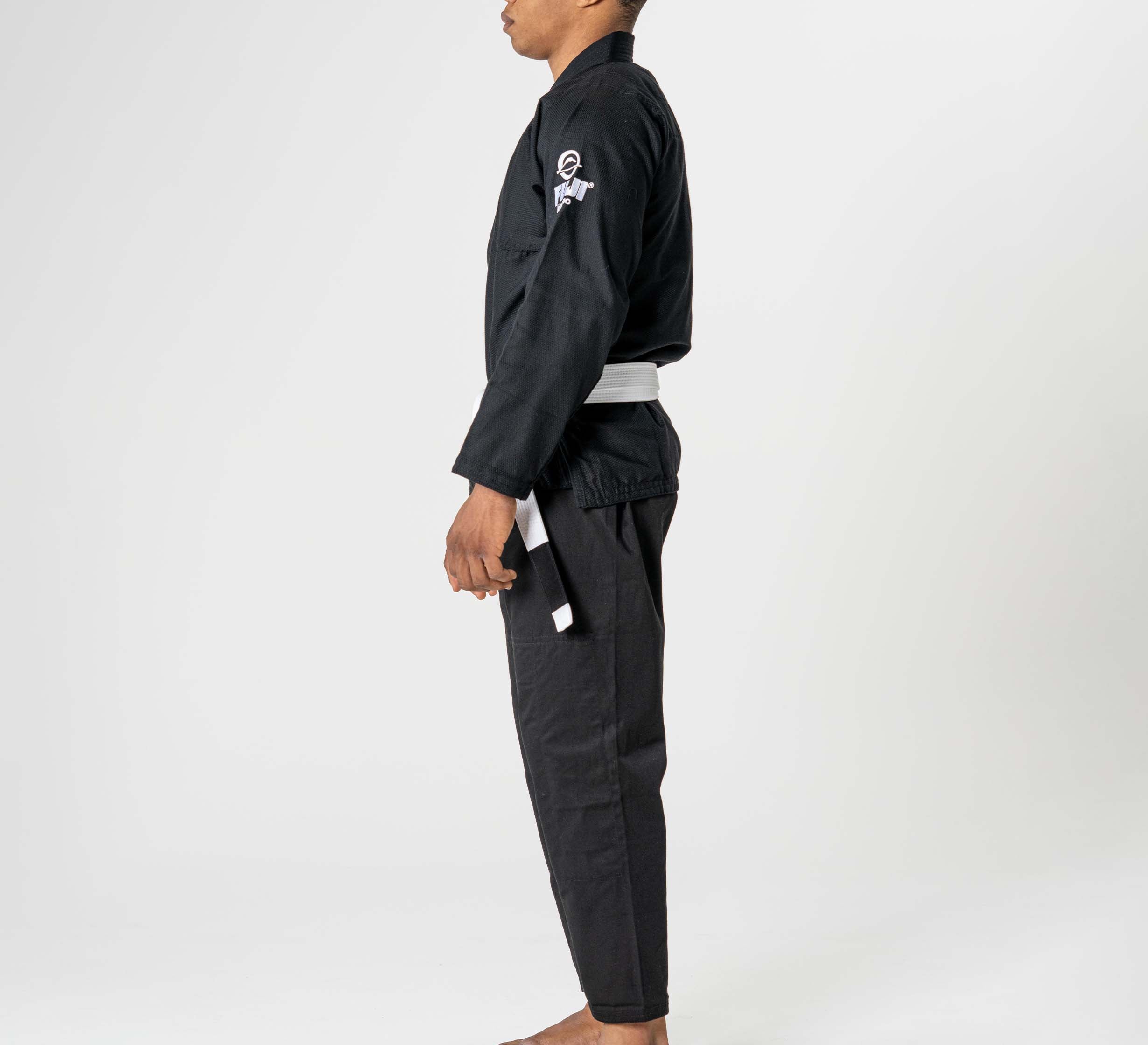 A person stands barefoot in profile wearing the Starter Adult BJJ Gi Black with a white belt against a plain white background.