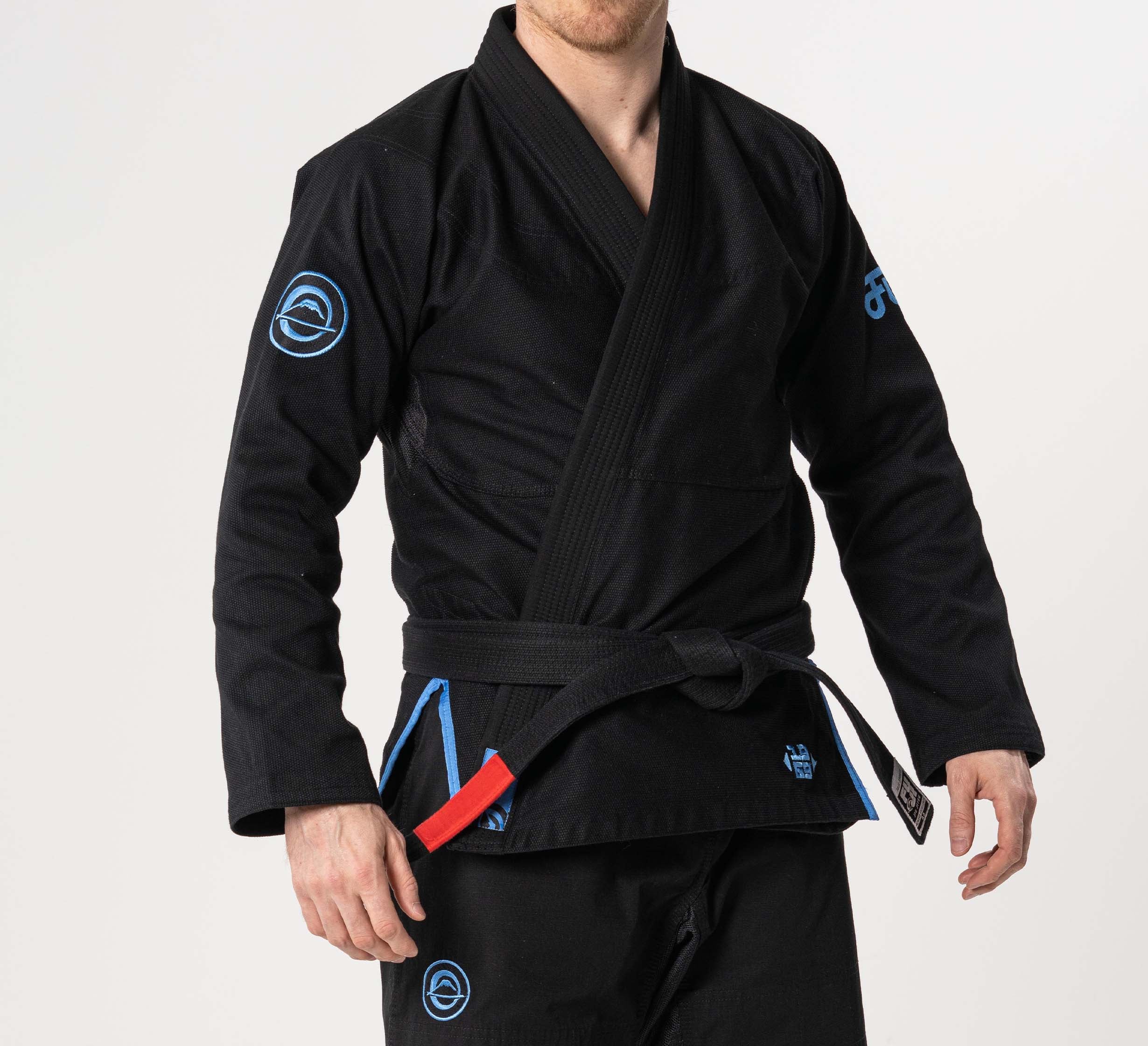 A person is wearing the Flow-Tech BJJ Gi in black with blue and grey accents, paired with a black belt featuring a red bar, standing against a plain white background.