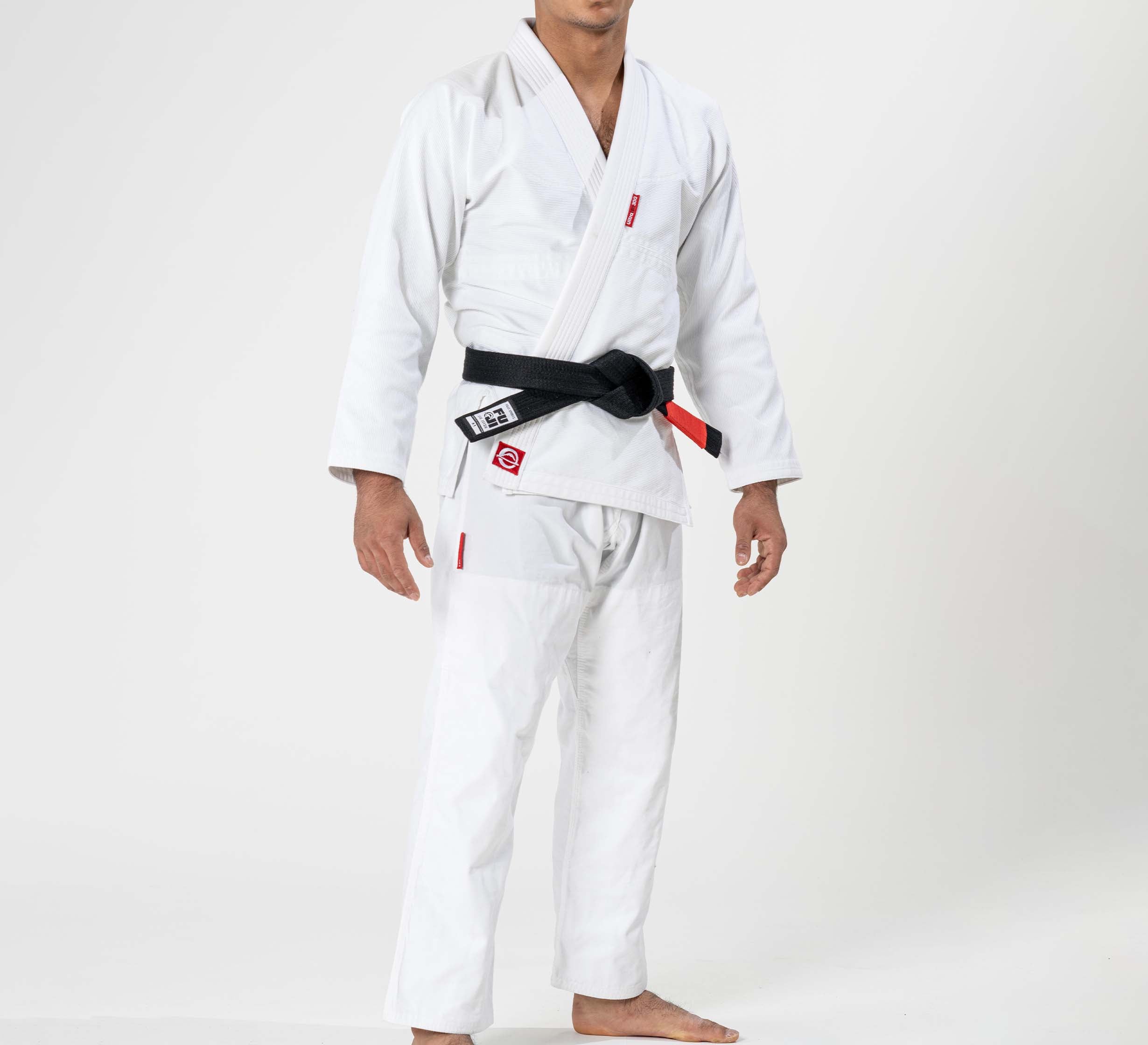 A person stands barefoot against a plain background, wearing the Ultra Lite 300 BJJ Gi White with a black belt at the waist. The person's head is not visible in the image.