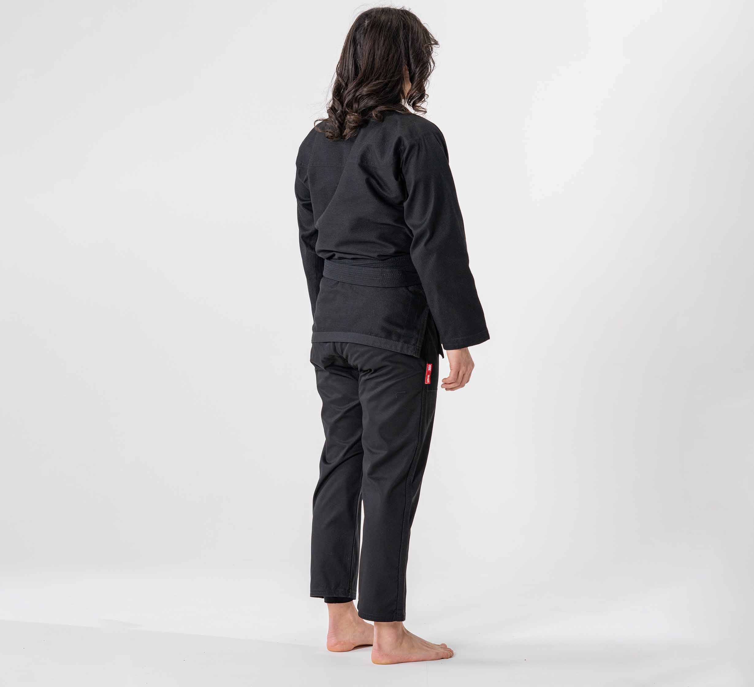 A woman with long hair, barefoot and wearing the Womens Ultra Lite 300 BJJ Gi Black, stands facing away from the camera against a plain white background.