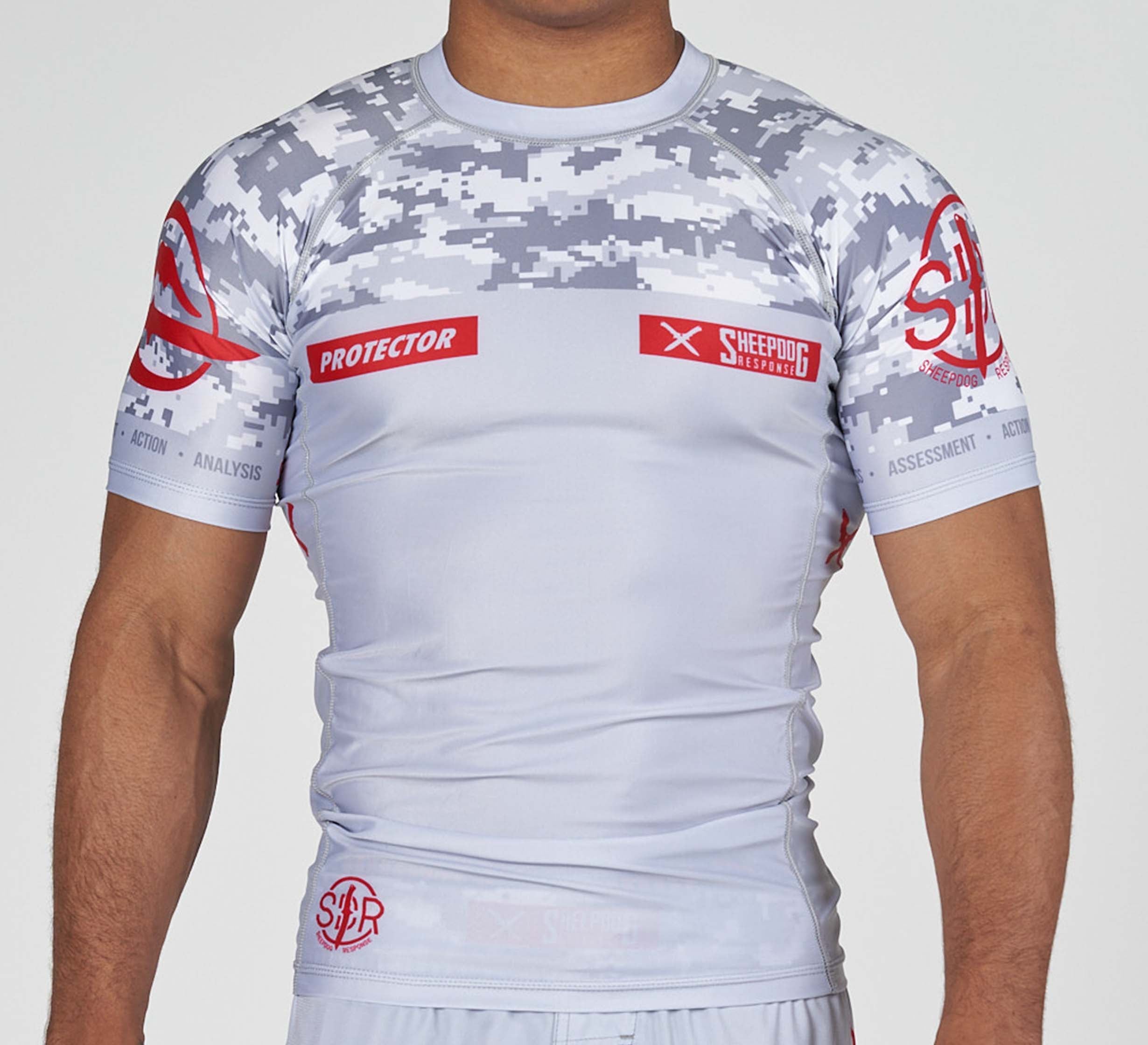 A man wears the Sheepdog Response Flex Lite Rashguard Grey, a fitted short-sleeve athletic shirt featuring gray digital camo on the shoulders and red PROTECTOR and SHEEPDOG RESPONSE logos on the chest.