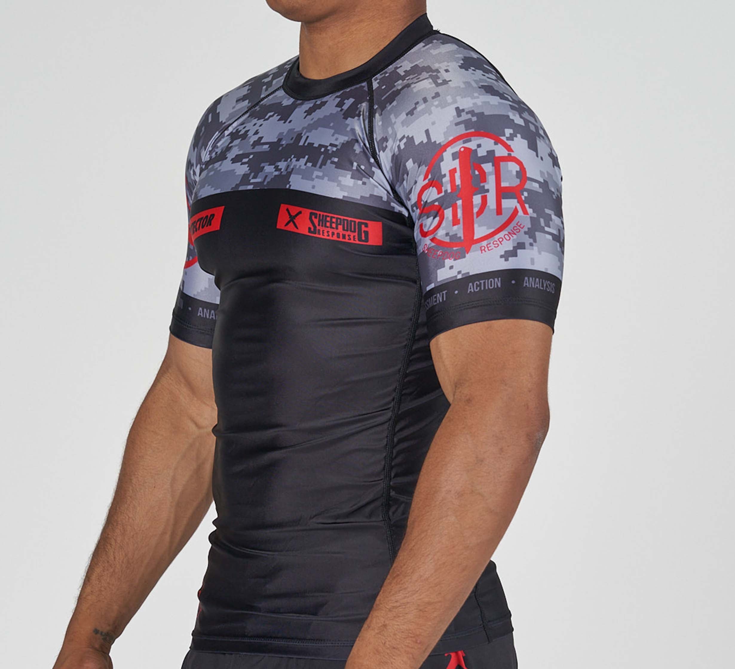 A man wearing the Sheepdog Response Flex Lite Rashguard Black, featuring a fitted black and gray camo design with red logos and text on the chest, shoulders, and arms, stands against a plain background.