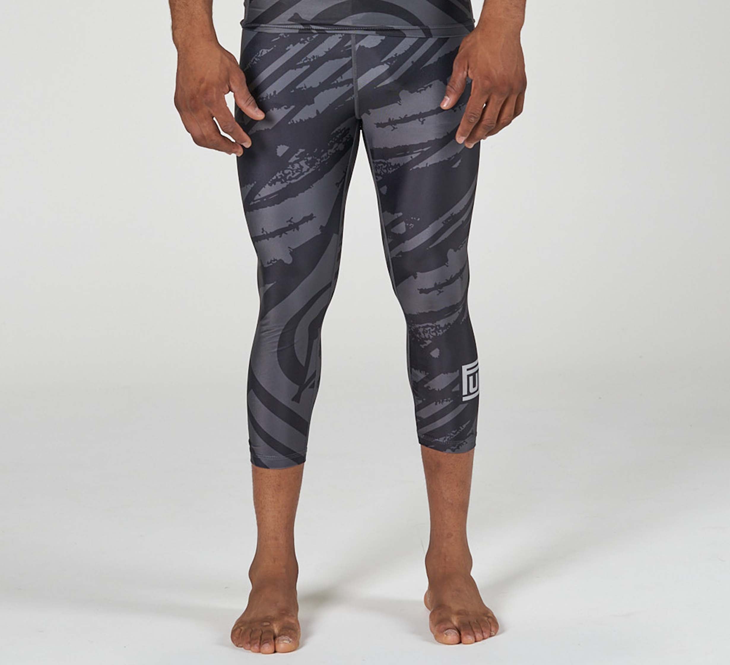 A person stands barefoot in Battle Flex Lite Spat Black leggings, which feature black and gray abstract diagonal patterns, a small white logo near the knee, and a light-colored plain background.