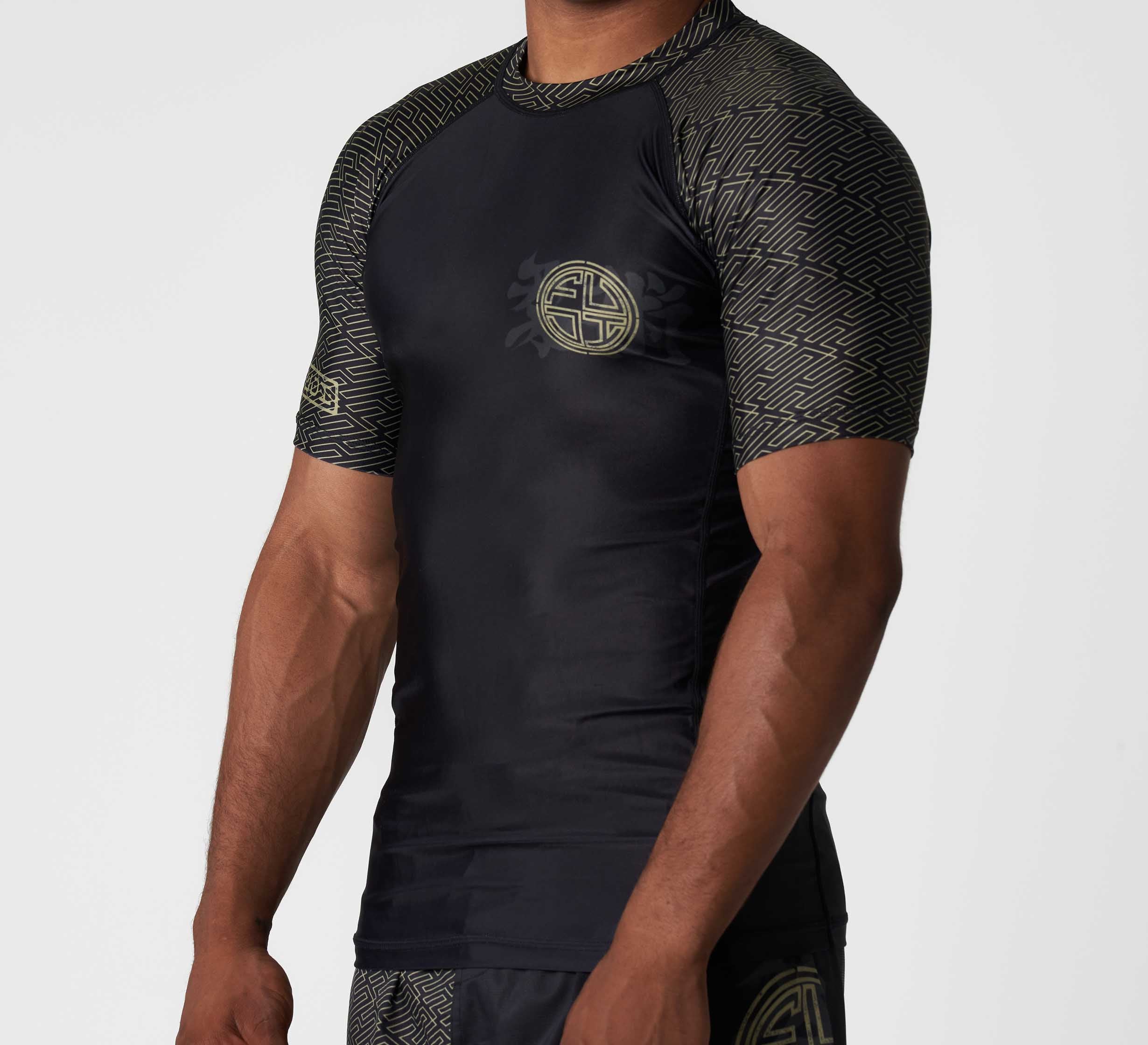 A person wearing the Bushido Flex Lite Rashguard—a black, short-sleeve top with gold geometric patterns on the shoulders, sleeves, and sides, plus a gold circular emblem on the chest and left leg of the shorts.