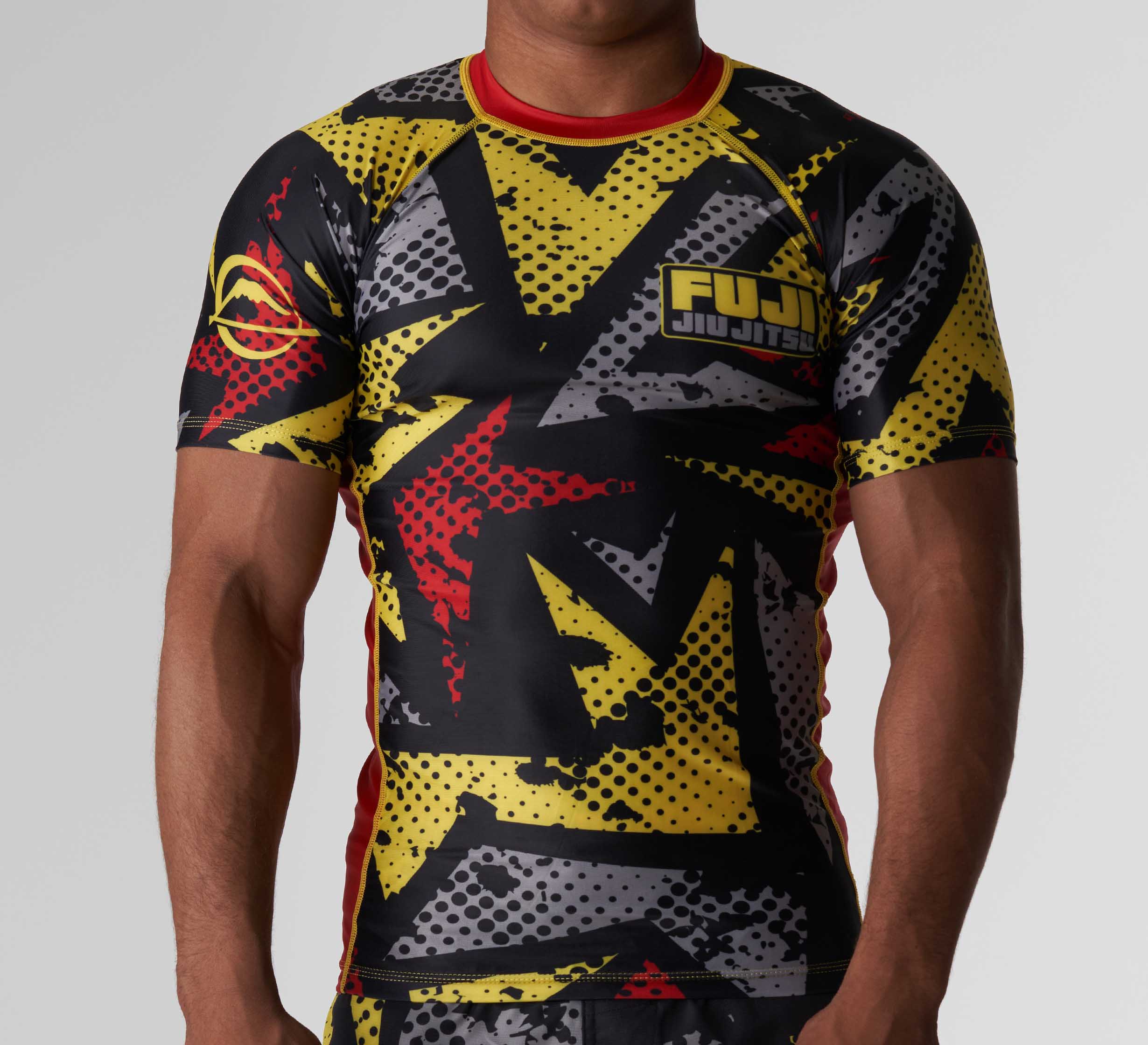 A person wearing the Shuriken Flex Lite Rashguard Black/Yellow, featuring short sleeves, an abstract black, yellow, and red graphic design, and the FUJI logo on the chest.