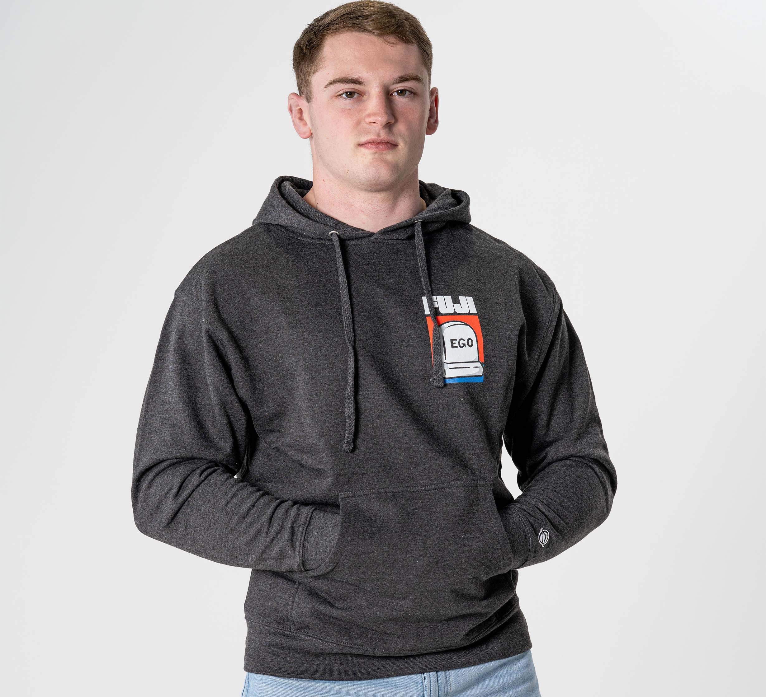 A young man stands facing the camera in the Submit Your Ego Hoodie Heather Black, featuring a colorful "EGO" graphic on the chest. He has short hair, hands in the front pocket, and poses against a plain white background.