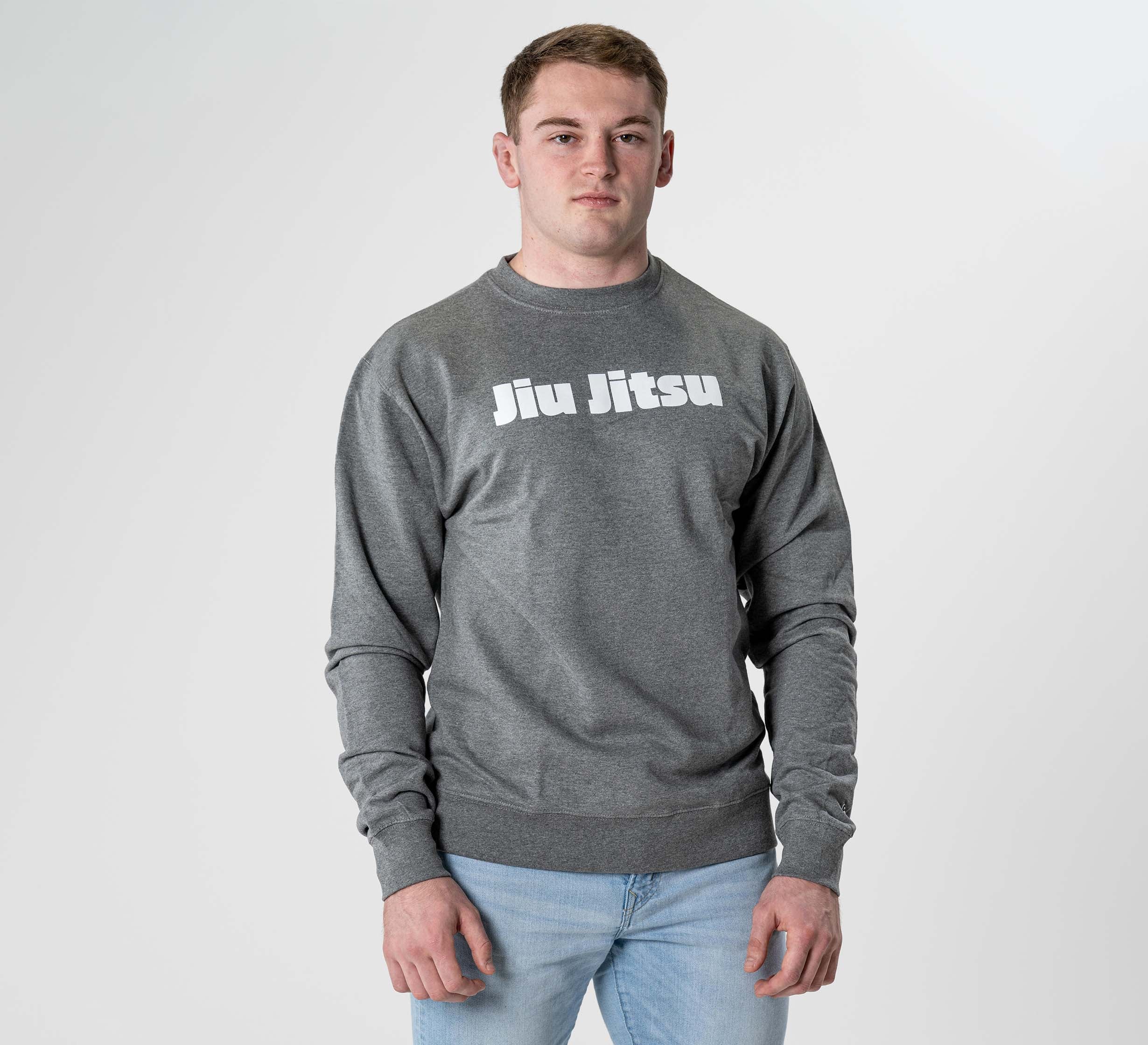 A person stands against a plain light background, wearing the Jiu Jitsu Player Crewneck Heather Grey sweatshirt with bold white "Jiu Jitsu" lettering and light blue jeans.