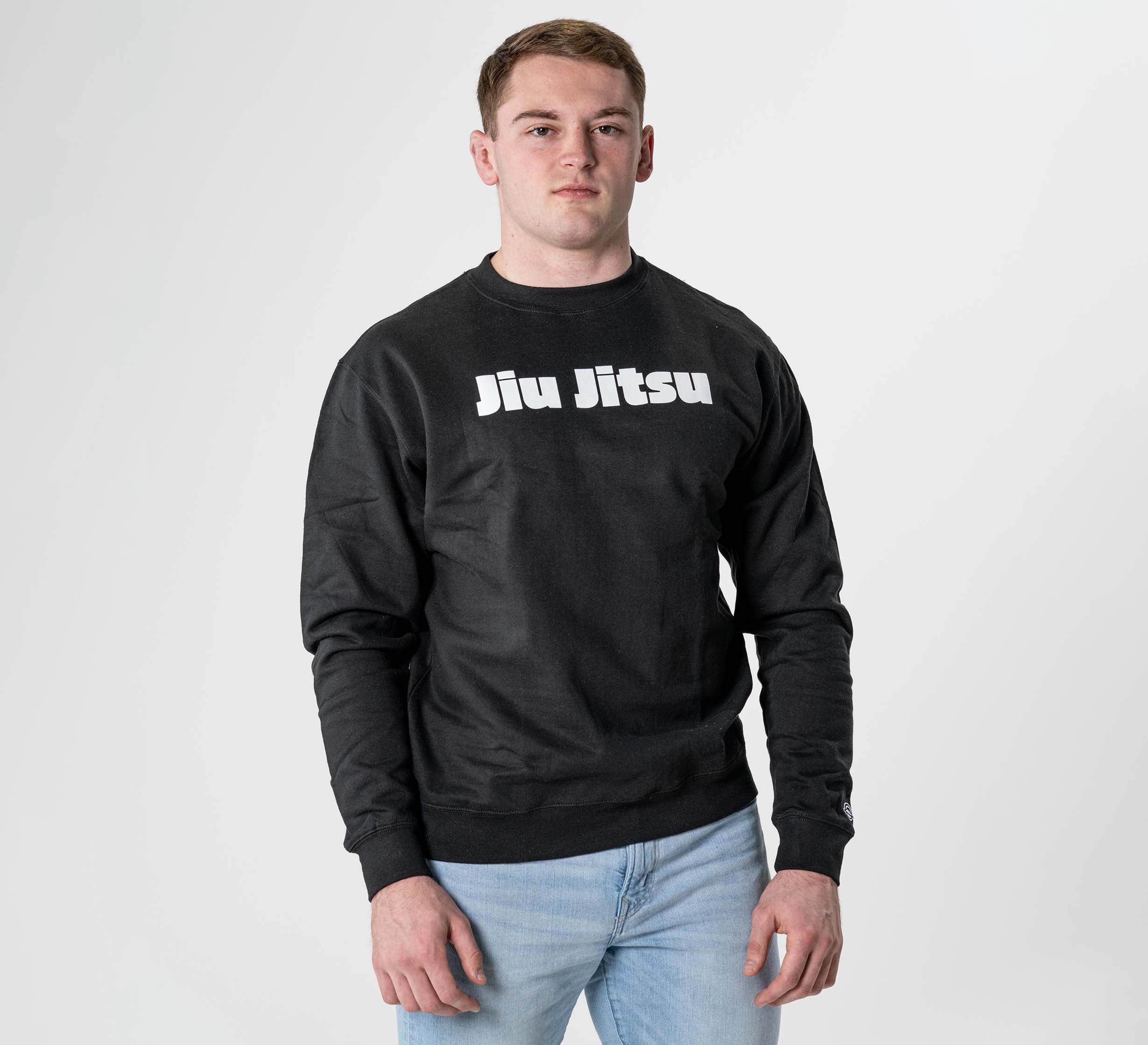 A young man poses against a plain white background, wearing the Jiu Jitsu Player Crewneck Black with bold white "Jiu Jitsu" lettering on the front and light blue jeans.