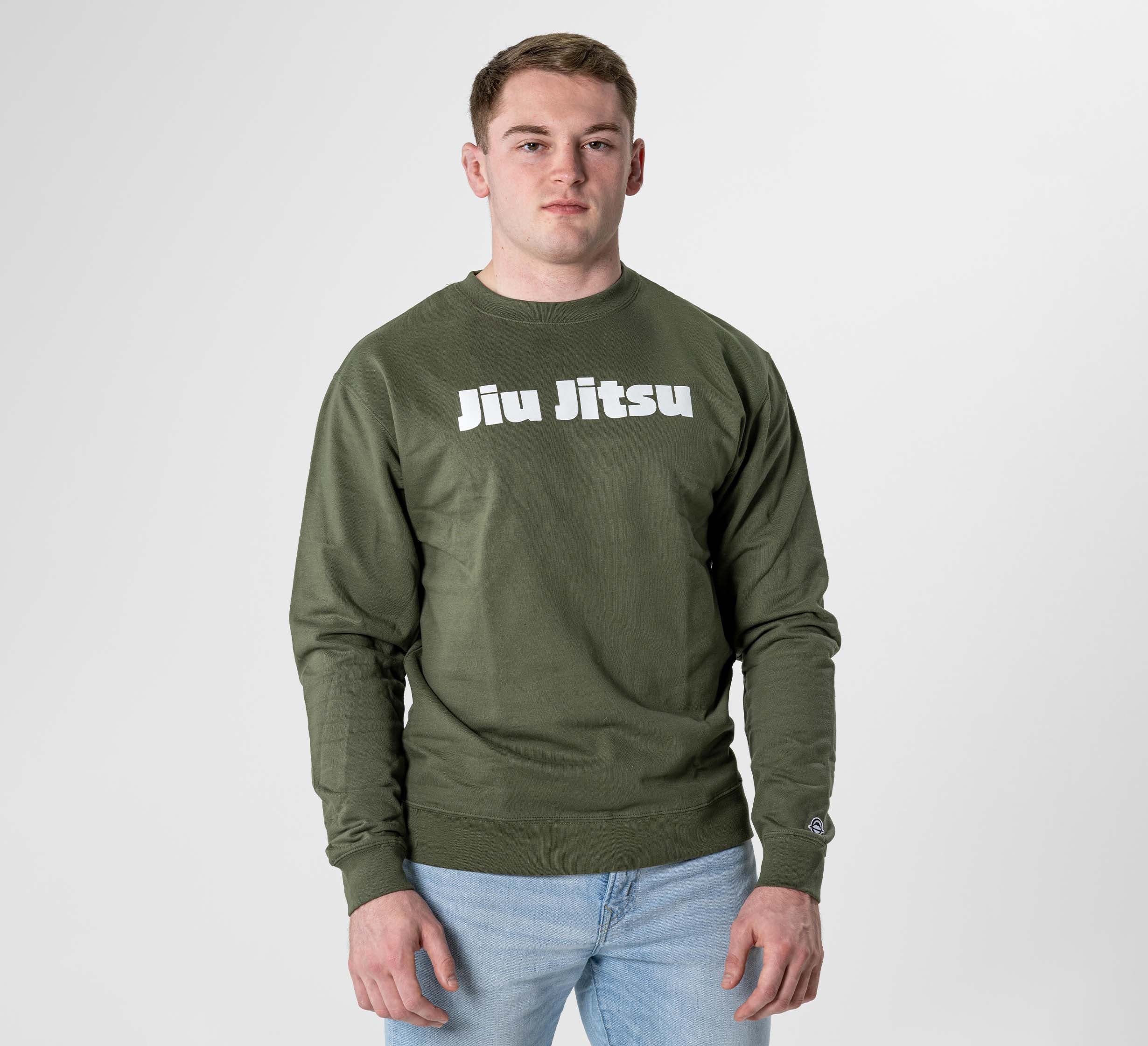 A young man with short hair stands against a plain light background, looking at the camera, wearing the Jiu Jitsu Player Crewneck in Military Green with bold white "Jiu Jitsu" lettering on the front.