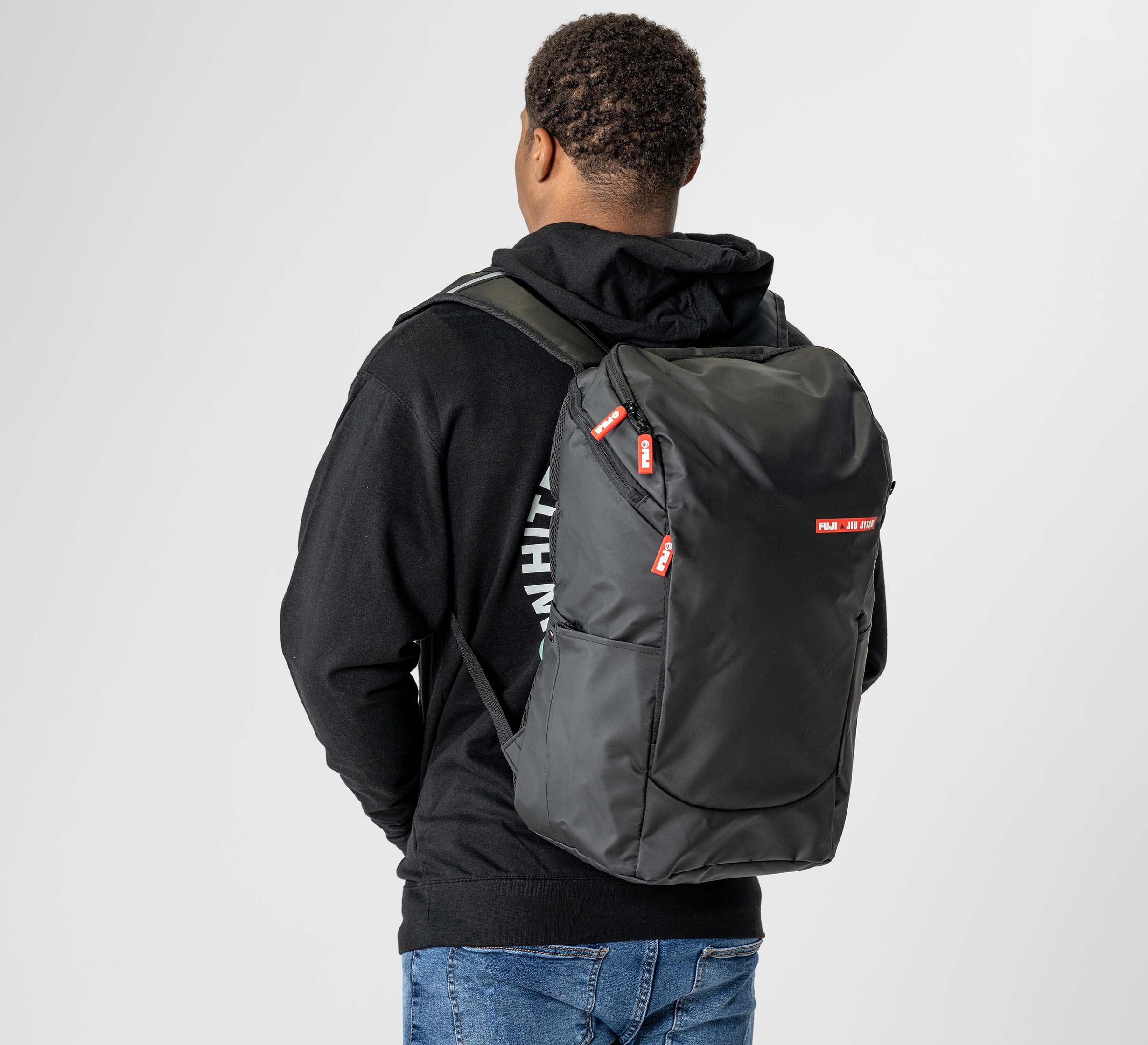 A person in a black hoodie and blue jeans is seen from behind carrying the Urban Day Backpack Black, featuring red accents and labels, against a plain, light-colored background.