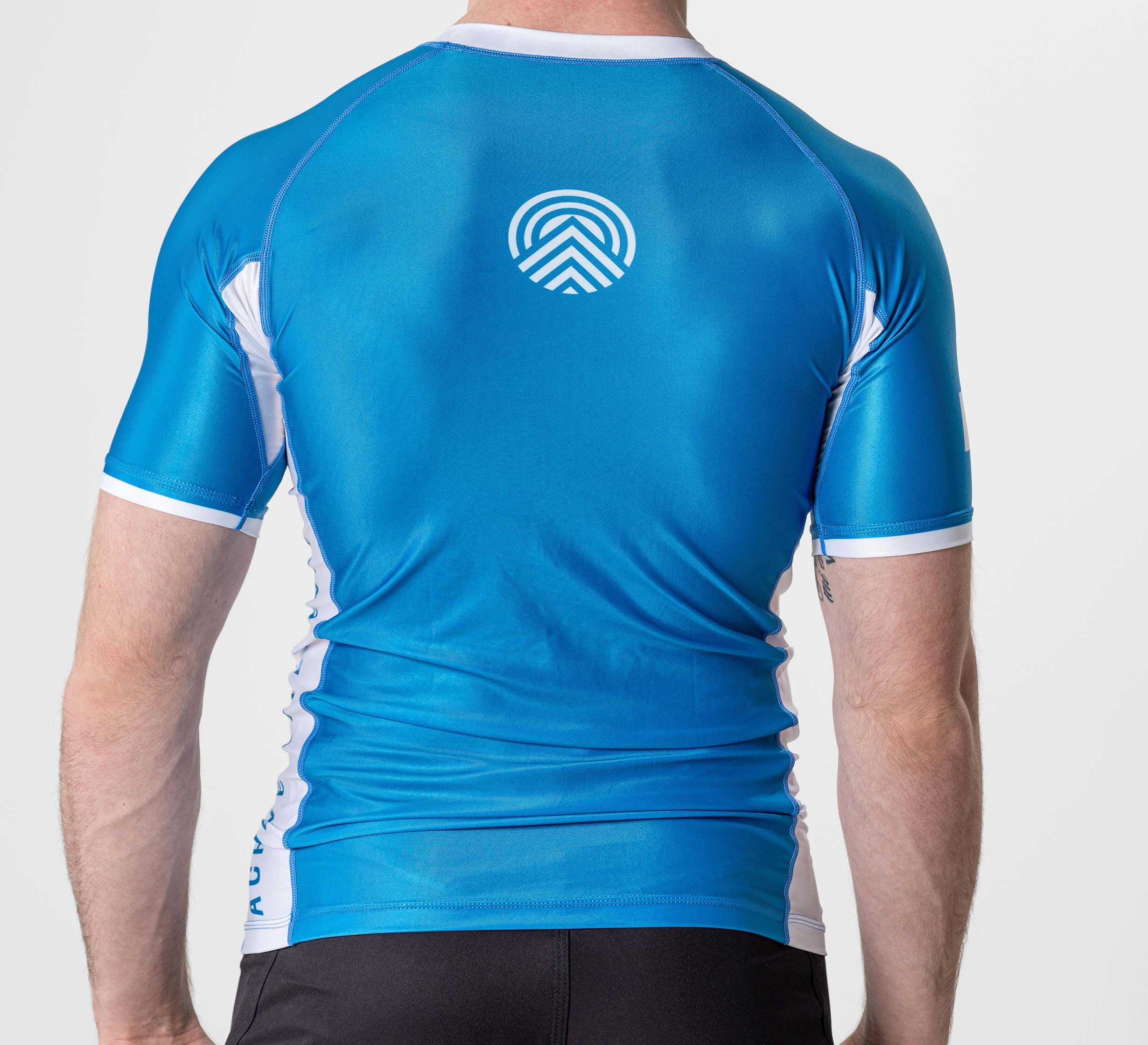 A person is shown from the back wearing the BJJ Flex Lite Rashguard Blue with short sleeves, a fitted design, and a white geometric logo on the upper center, paired with black shorts.