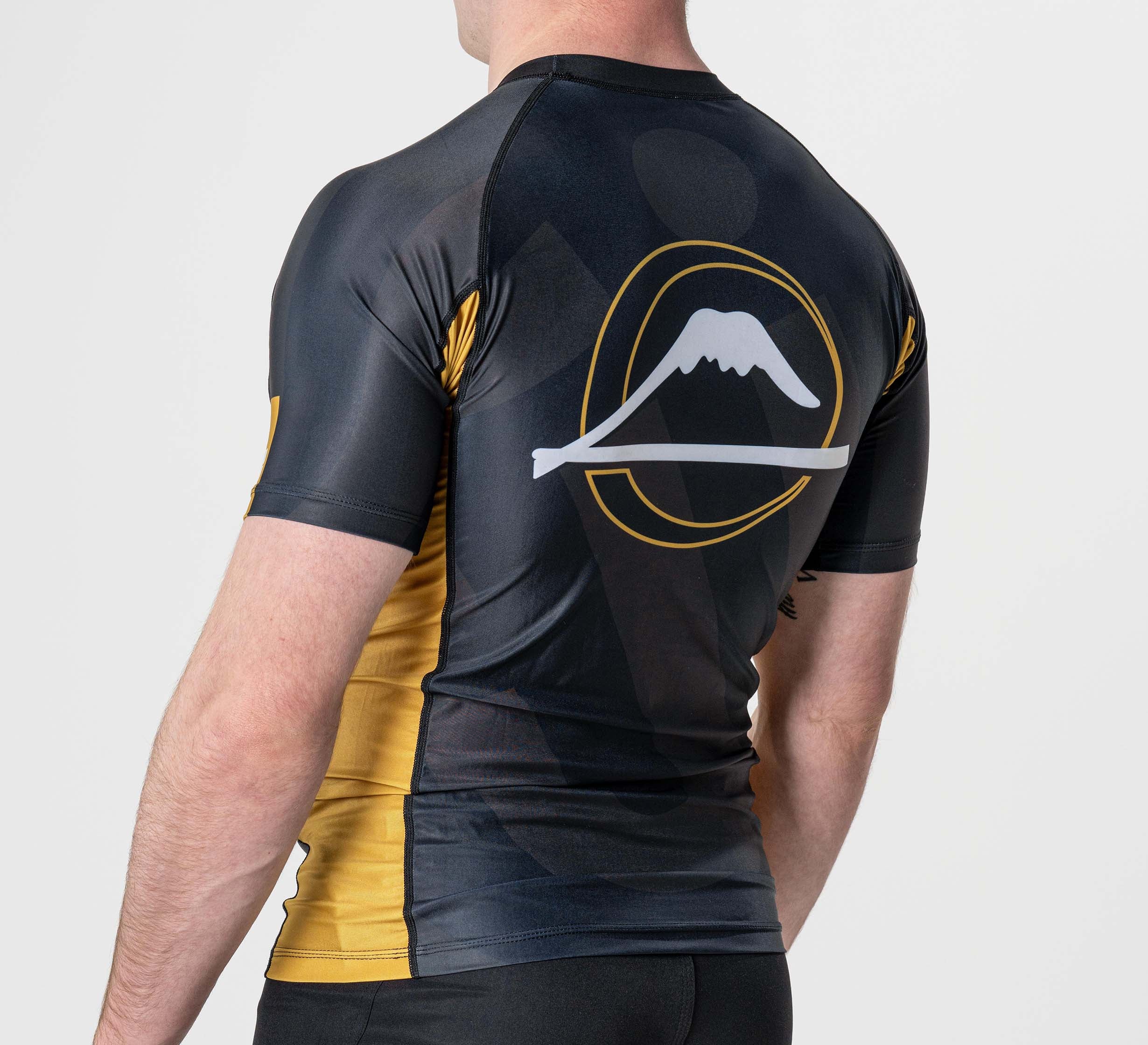 A person wearing the Rebel Flex Lite Rashguard Black, a short-sleeved athletic shirt featuring a white mountain design and yellow circular accents on the back, stands against a plain light background.