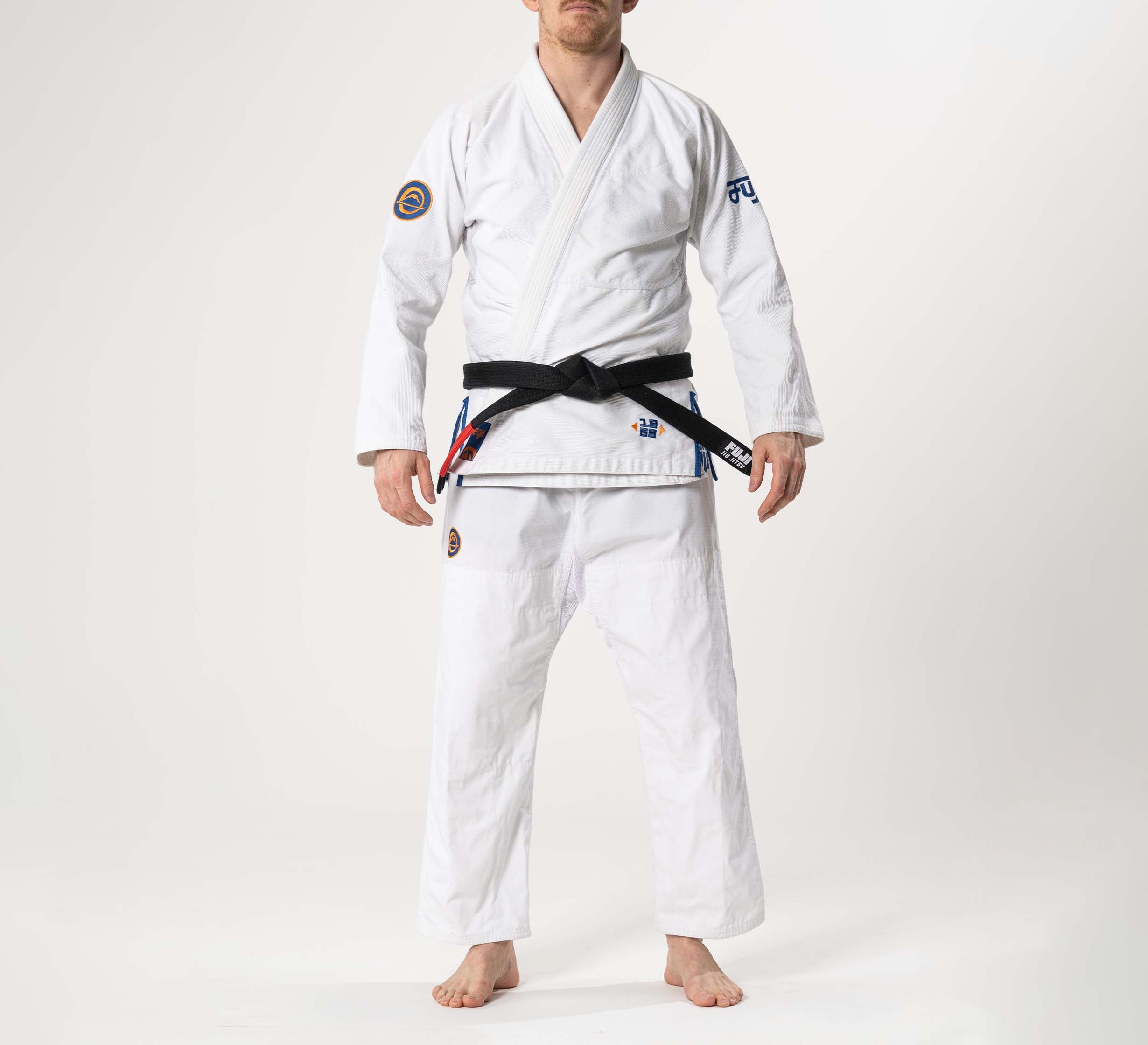 A person stands barefoot on a white background, wearing the Flow-Tech BJJ Gi in white/navy/orange and a black belt, arms relaxed at their sides. Their face is not visible.