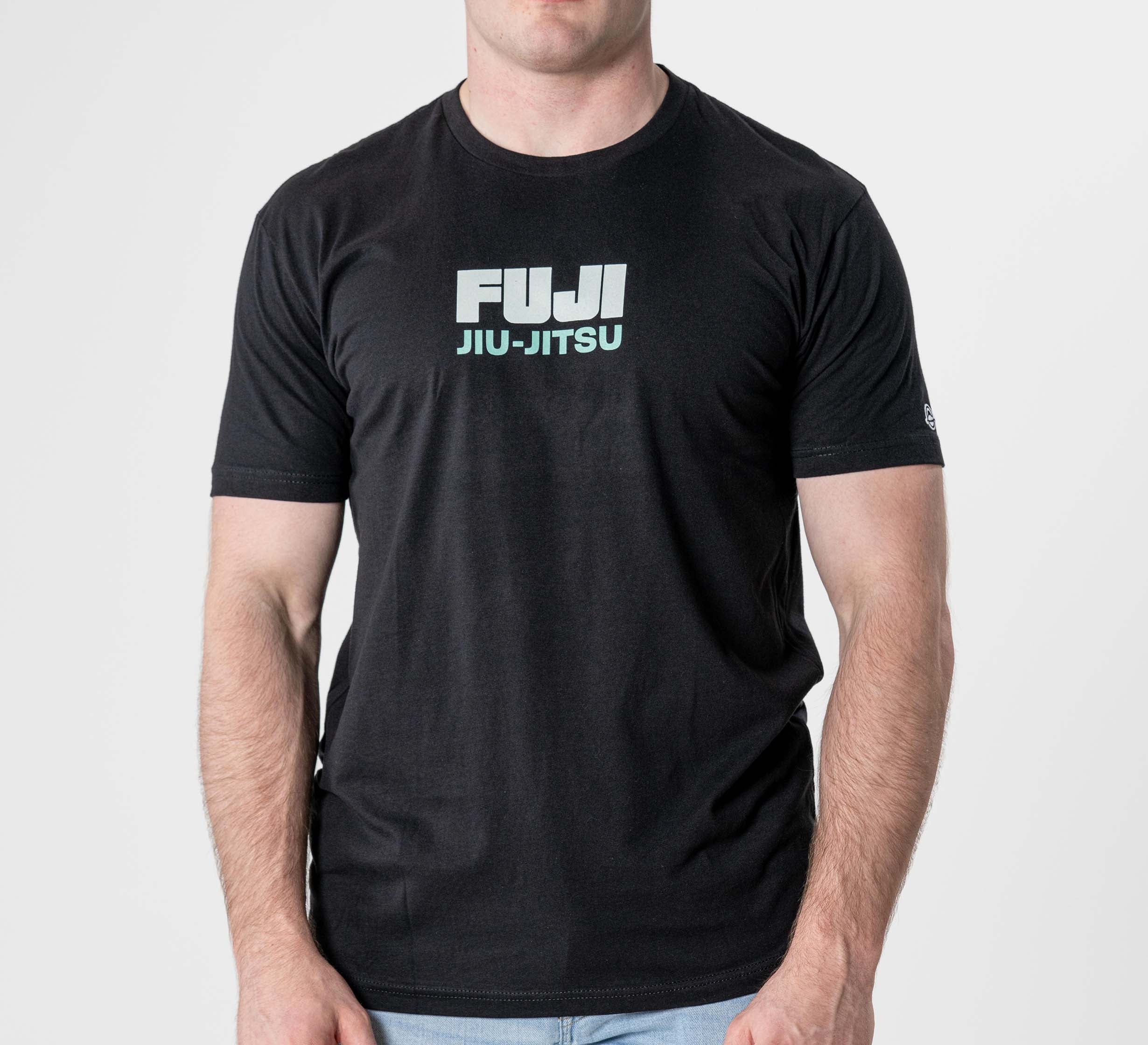 A man wearing the Black Belt Mentality T-Shirt Black, featuring bold white "FUJI JIU-JITSU" lettering on the front, stands against a plain light background.