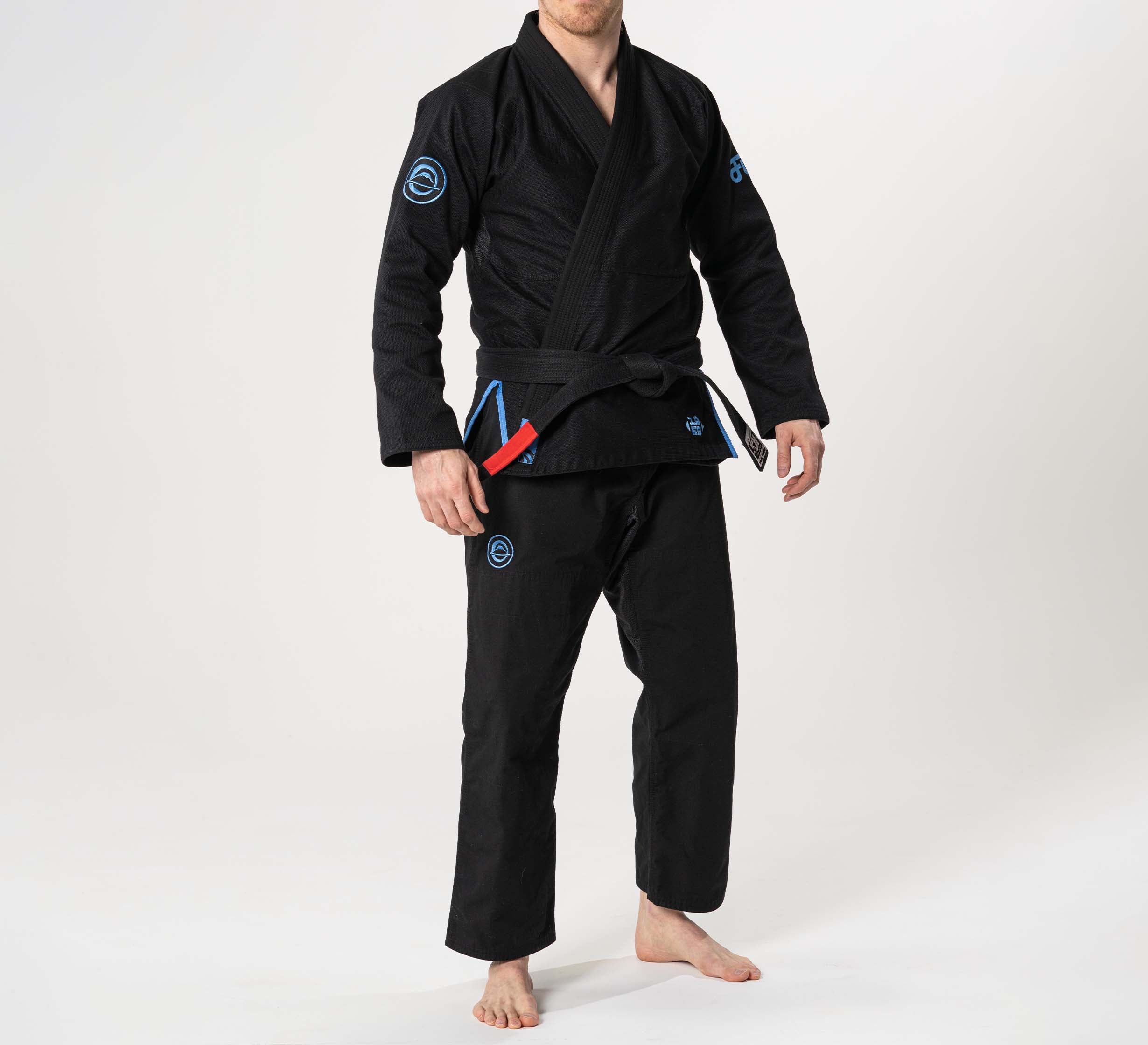 A person wears the Flow-Tech BJJ Gi in black with blue and grey accents, standing barefoot on a white background and holding a red belt stripe in their left hand. Their face is partially obscured.