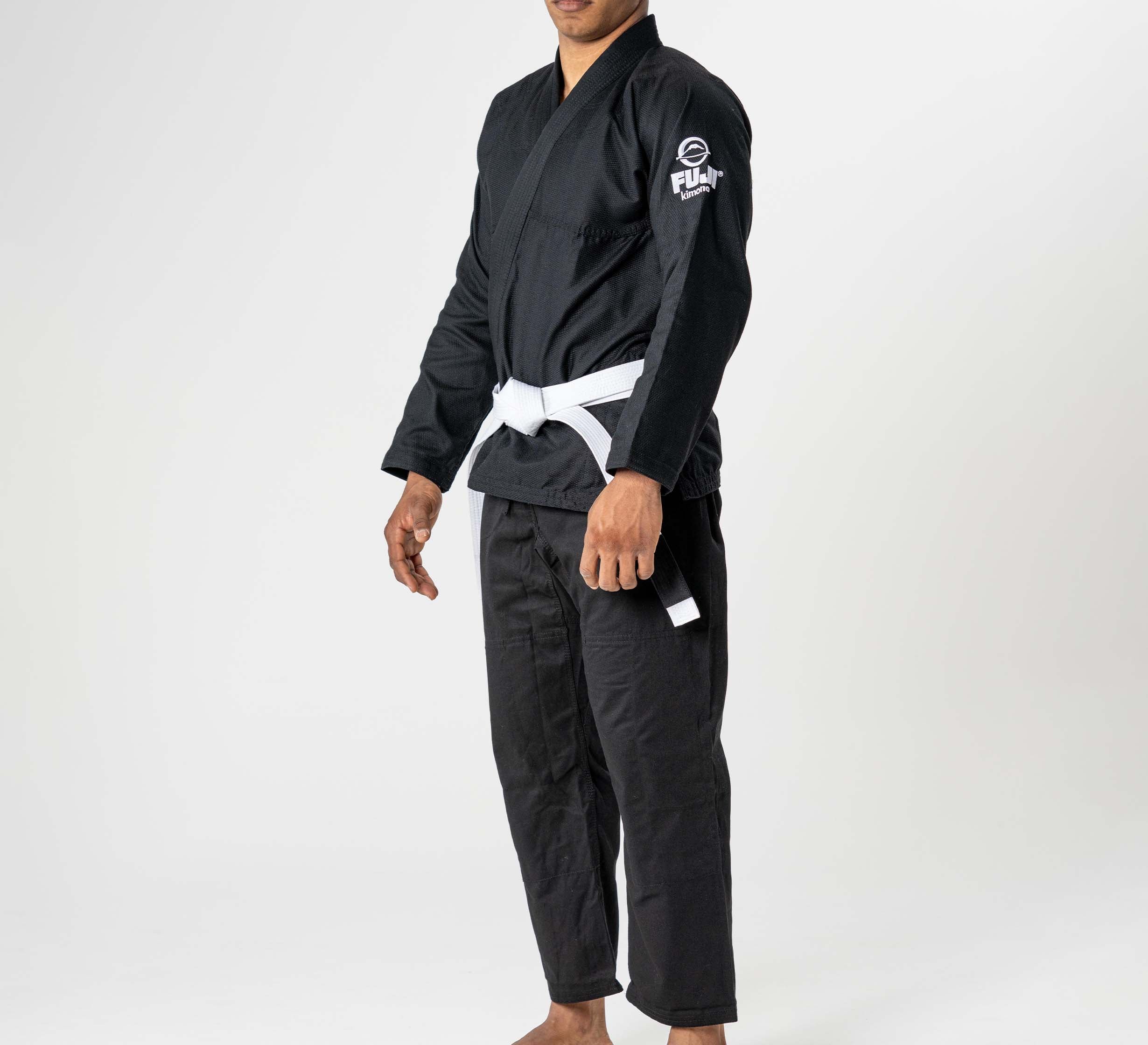 A person stands barefoot against a plain white background wearing the Starter Adult BJJ Gi Black with a white belt; the gi features a Fuji logo on the left sleeve. The person's face is partially out of frame.