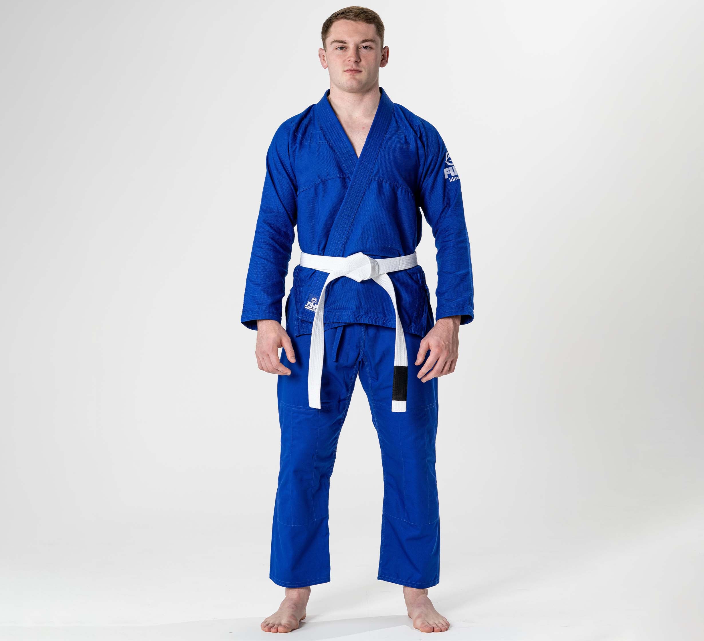 A person stands barefoot, facing forward, wearing the Starter Adult BJJ Gi Blue with a white belt, against a plain white background.