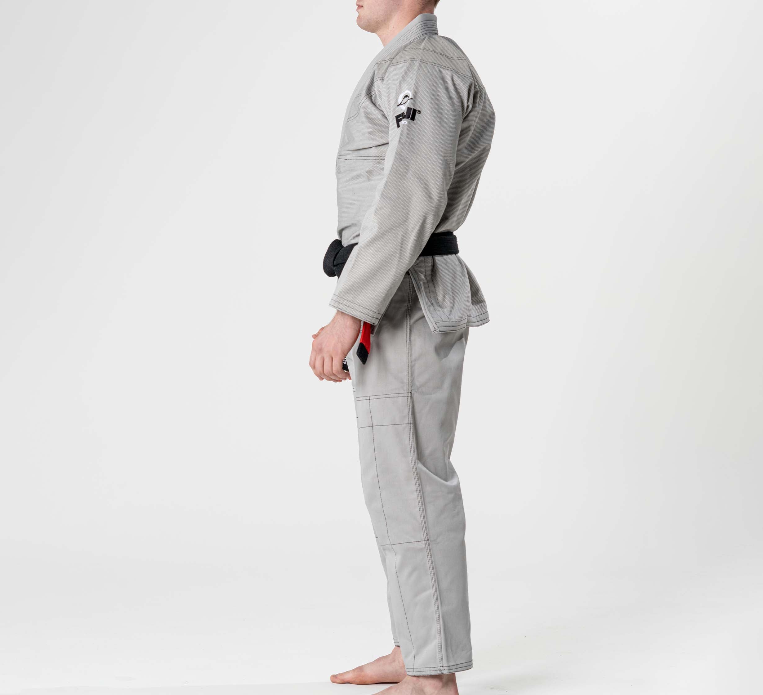 A person stands barefoot in profile on a white background, wearing the Lightweight BJJ Gi Grey with a black belt and an upper arm patch. The gi is light gray, and the face is partially out of frame.