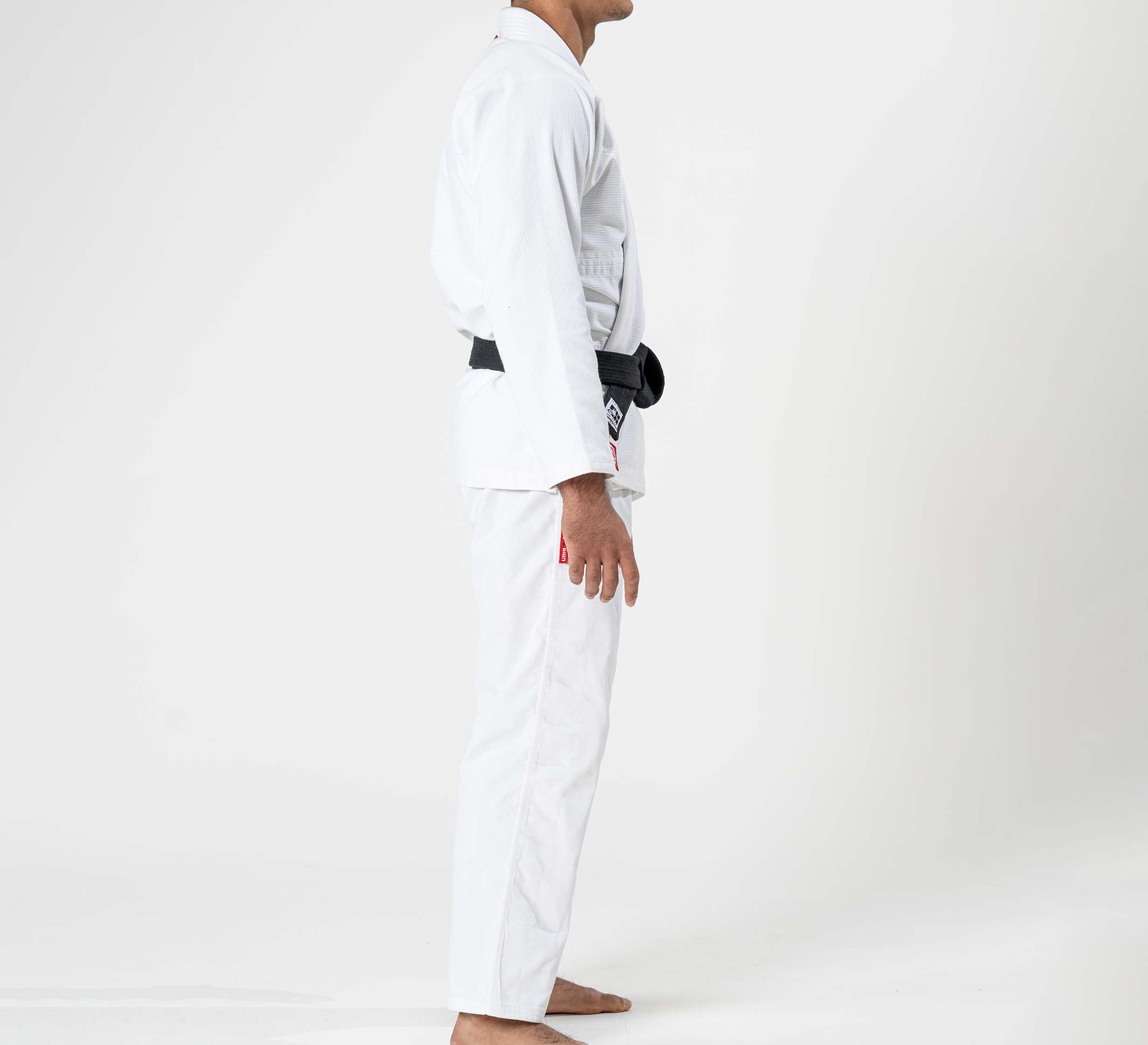 A person models the Ultra Lite 300 BJJ Gi White, wearing a black belt and standing barefoot in side profile against a plain white background.