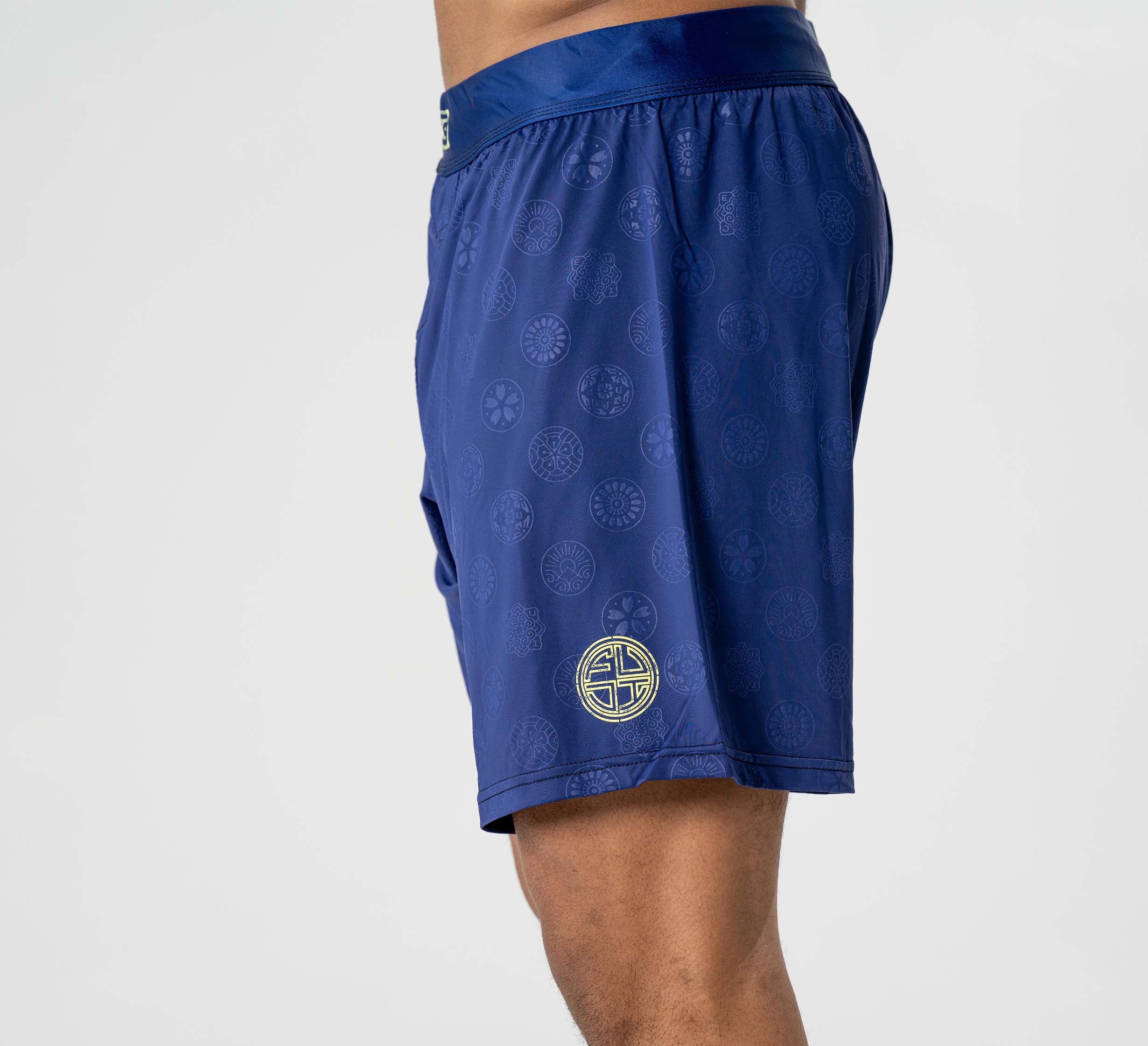 A person shown from the side wears Shogun Heat Gear Shorts in navy, featuring a subtle pattern and a gold circular emblem on the leg, against a plain light background.