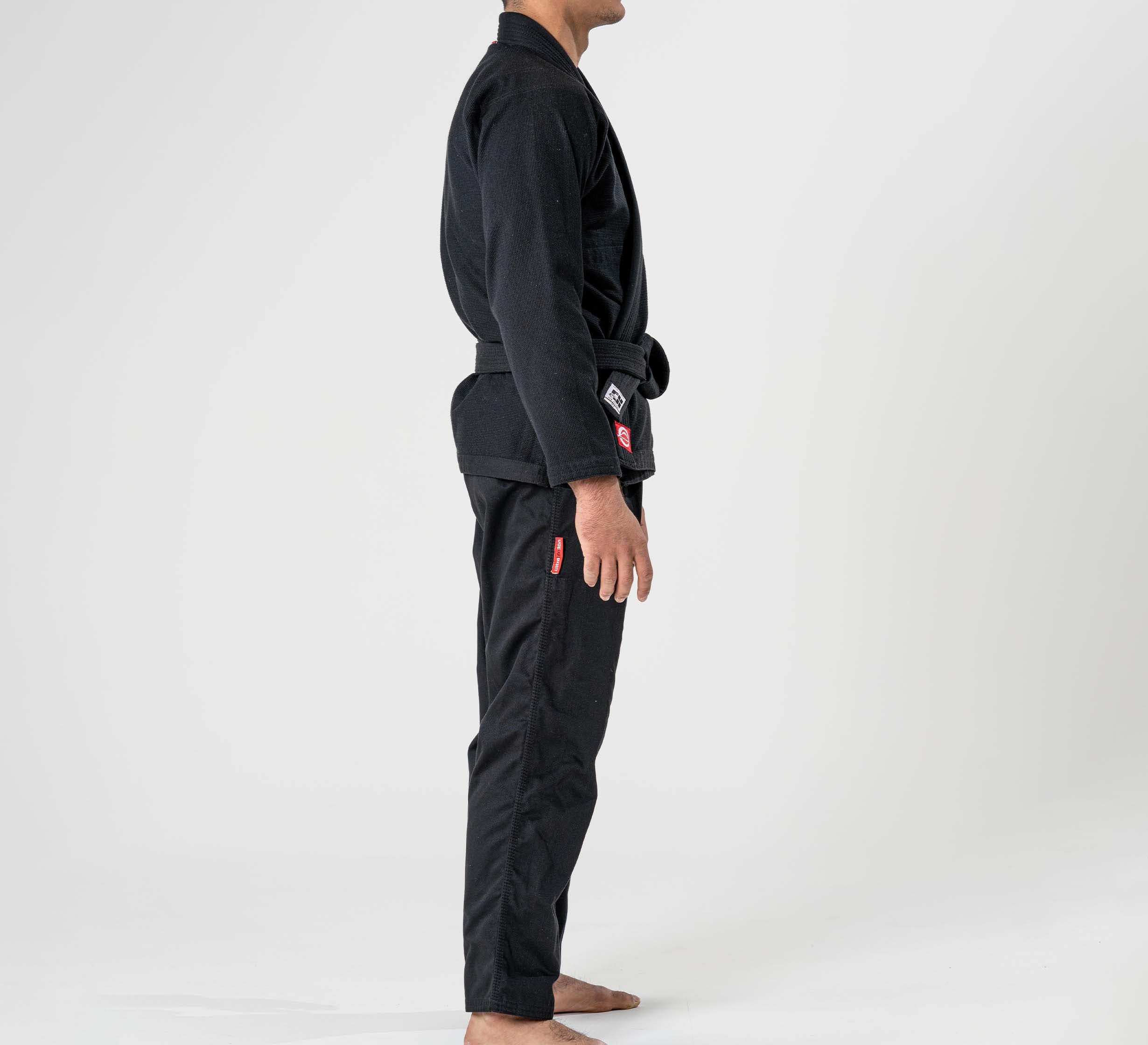 A person stands barefoot in a side profile wearing the Ultra Lite 300 BJJ Gi Black with a black belt, set against a plain white background.