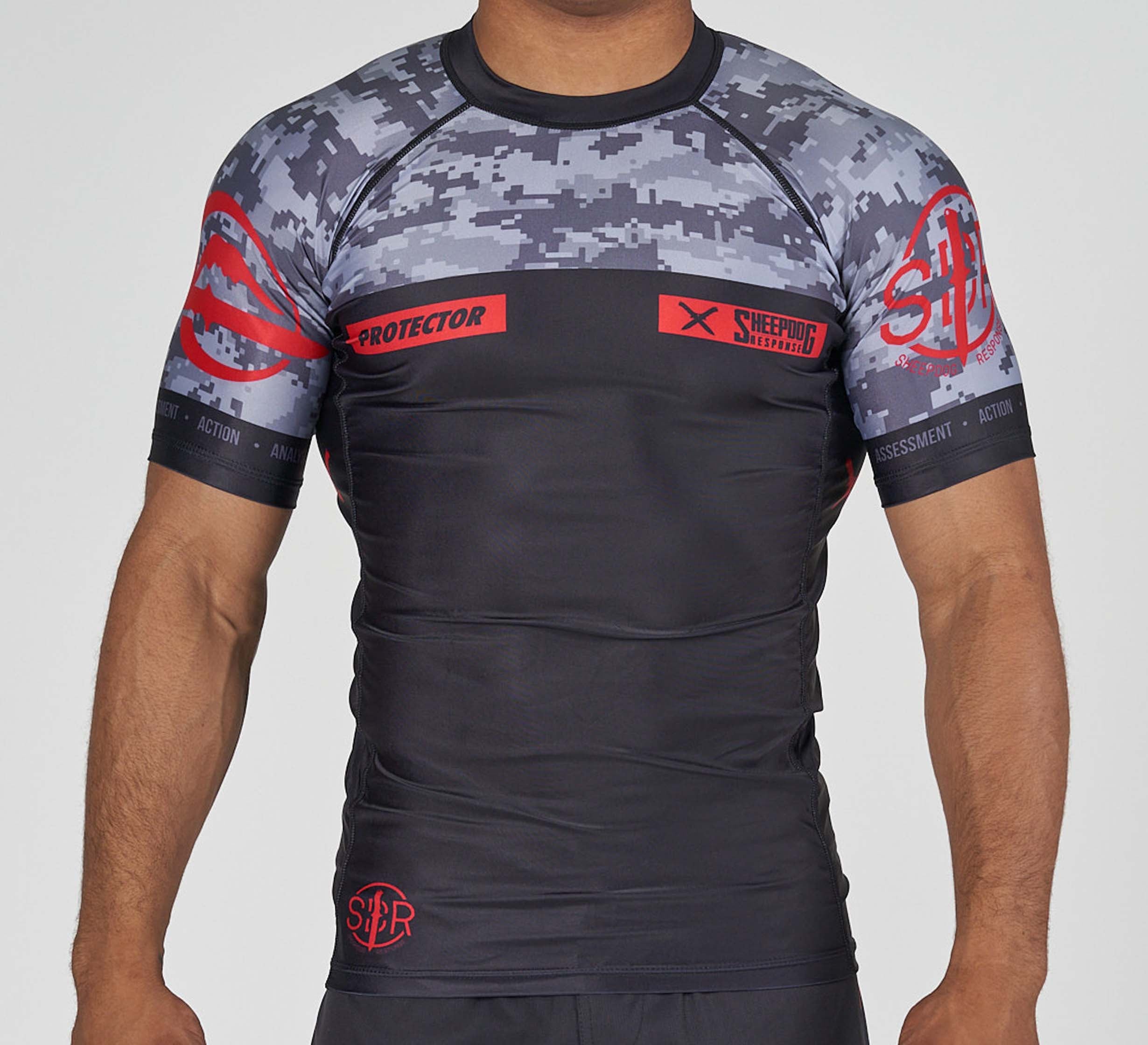A man wears the Sheepdog Response Flex Lite Rashguard in black and gray with red PROTECTOR and SHEPHERD logos, standing against a plain background.