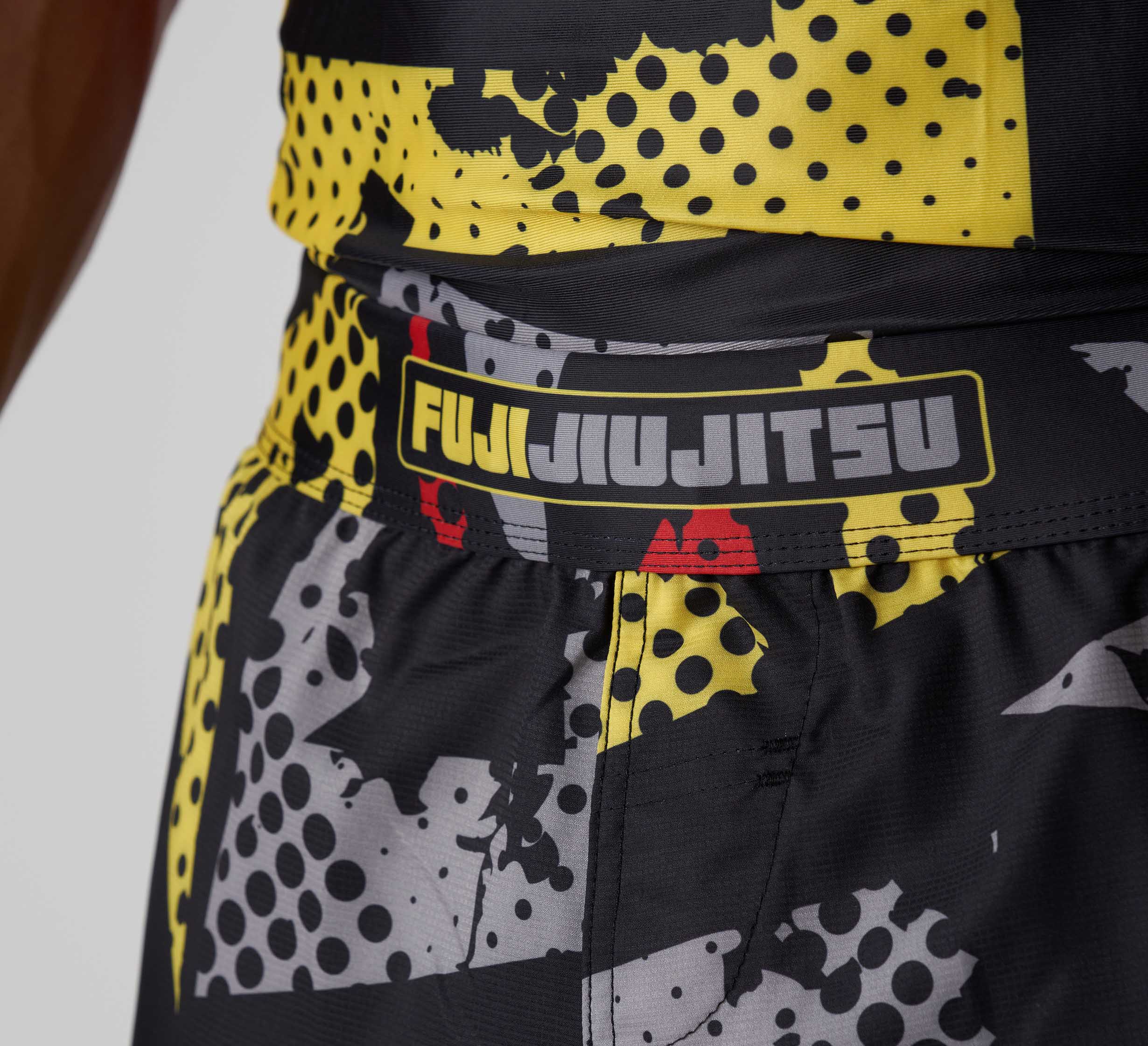 Close-up of a person's waist wearing Shuriken Flex Lite Shorts Black/Yellow, featuring a bold abstract pattern and FUJI JIU JITSU logo on the waistband.