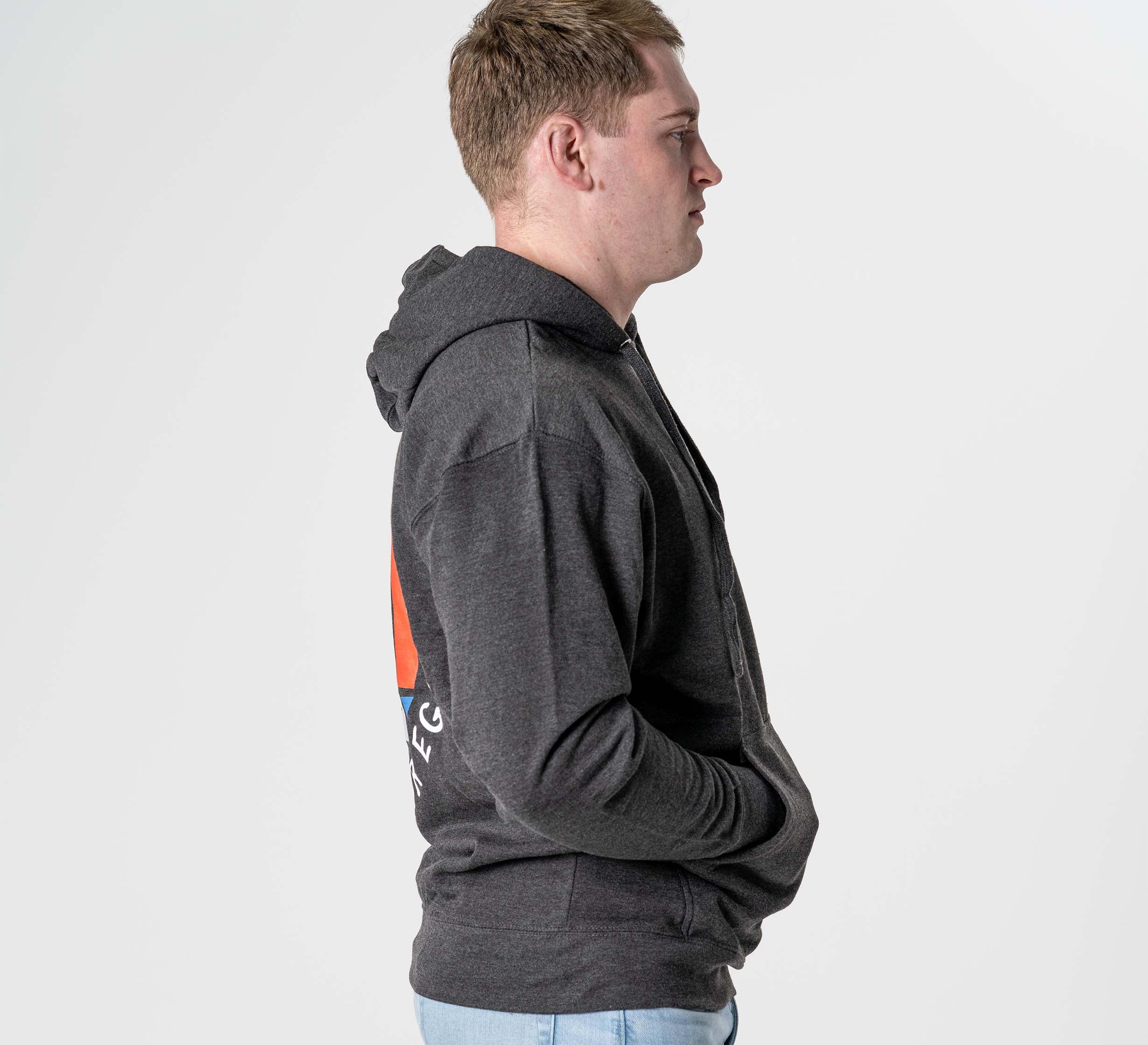 A young man wearing the Submit Your Ego Hoodie in Heather Black and light blue jeans stands in profile against a white background, hands in his pockets and looking straight ahead.
