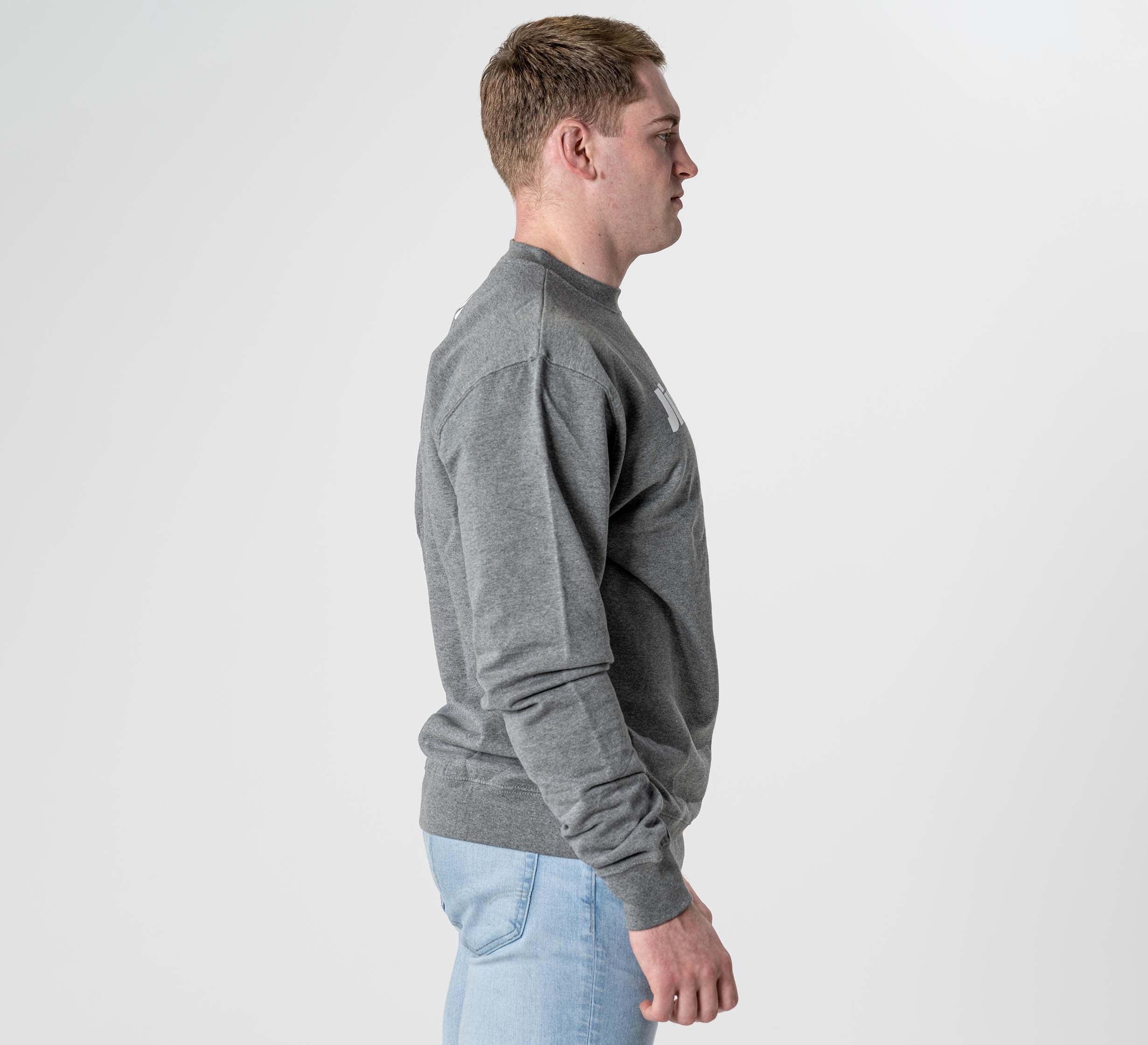 A man stands in profile facing right, wearing the Jiu Jitsu Player Crewneck Heather Grey and light blue jeans against a plain white background.