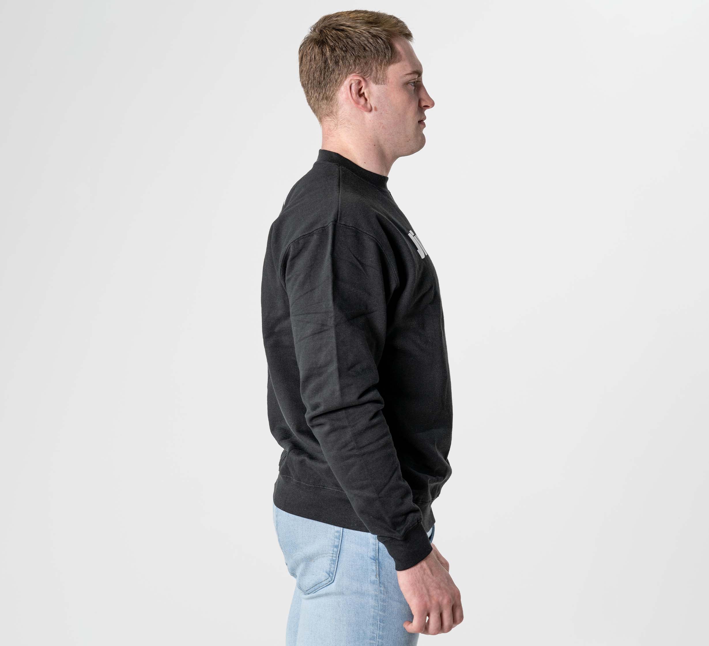 A man with short light brown hair stands in profile facing right, wearing the Jiu Jitsu Player Crewneck Black and light blue jeans against a plain white background.