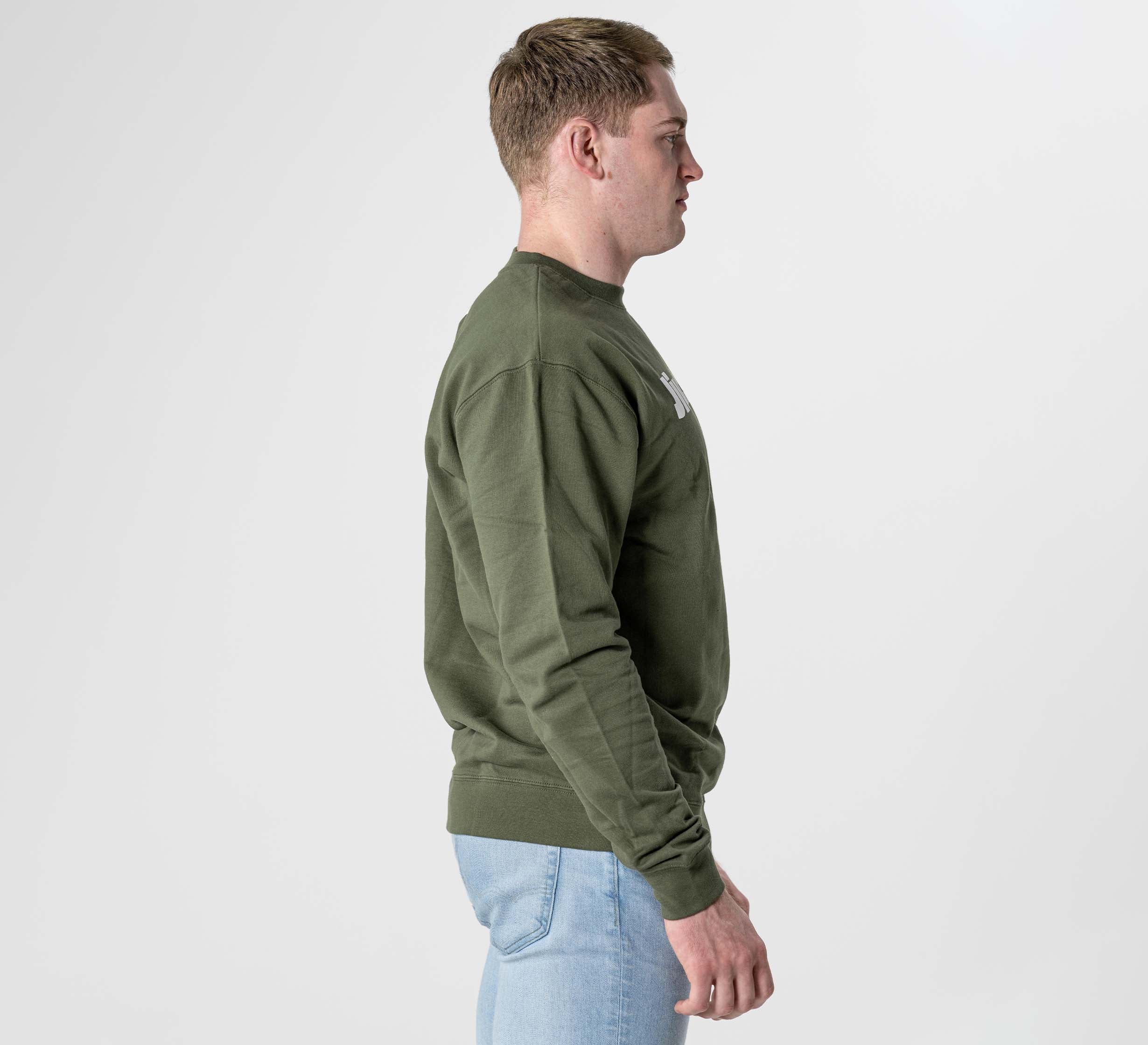 A man in profile faces right, wearing the Jiu Jitsu Player Crewneck in military green with light blue jeans, standing against a plain white background.