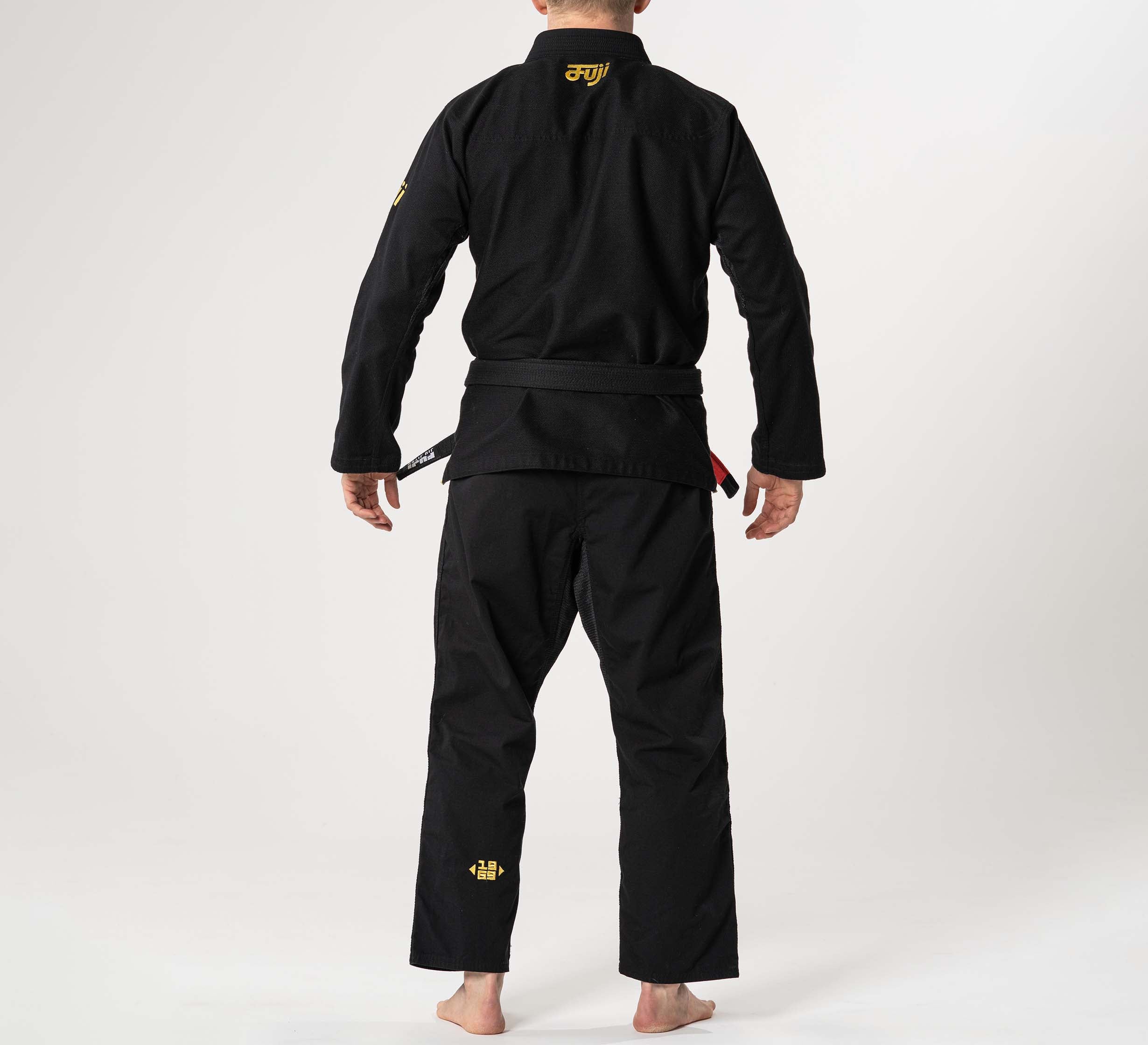 A person wearing the Flow-Tech BJJ Gi Black/Gold stands barefoot, facing away against a white background. Yellow embroidery accents are visible on the gi's upper back and pants.