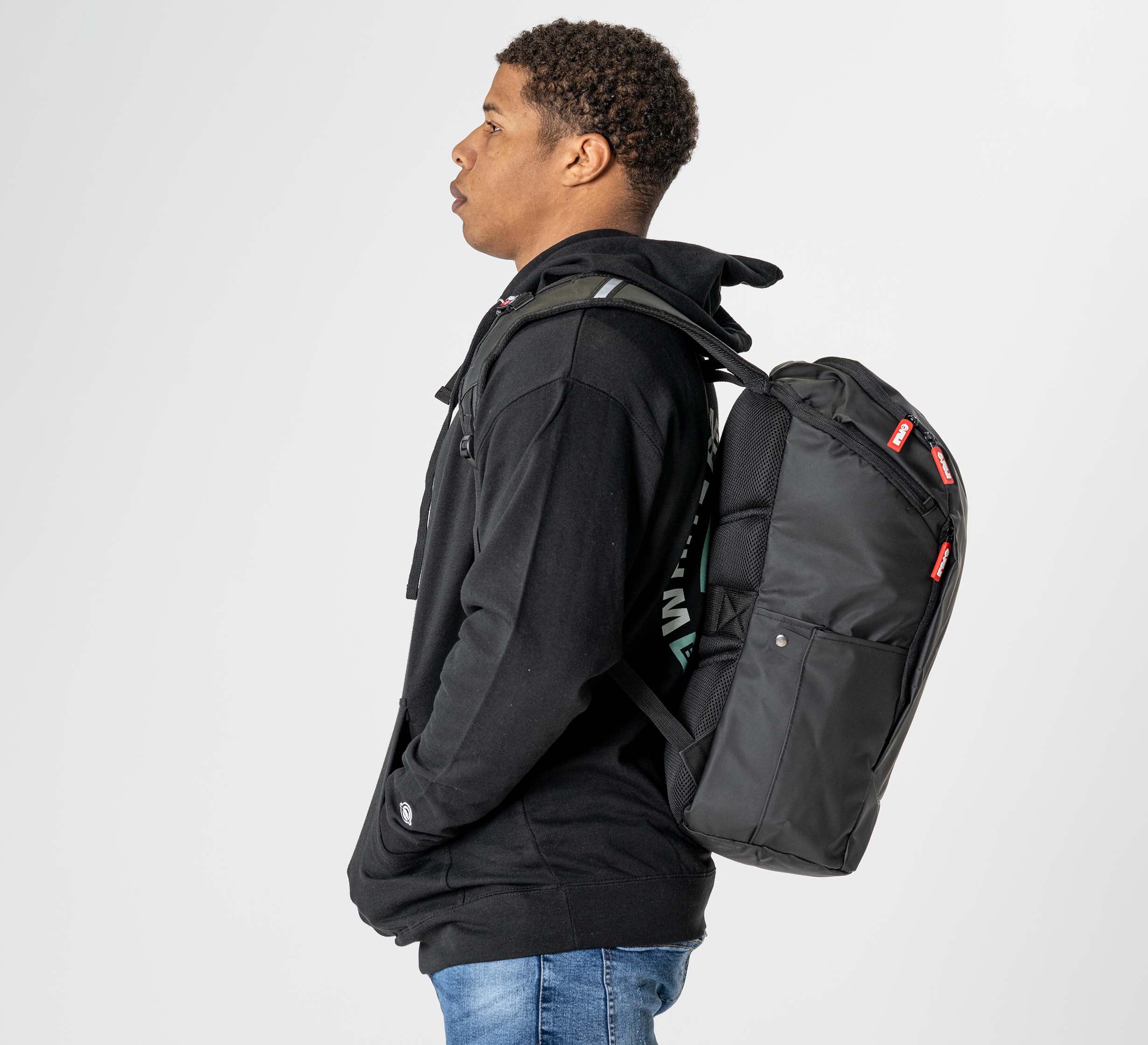 A person in a black hoodie and blue jeans stands sideways, wearing the Urban Day Backpack Black with red tags, against a plain white background.