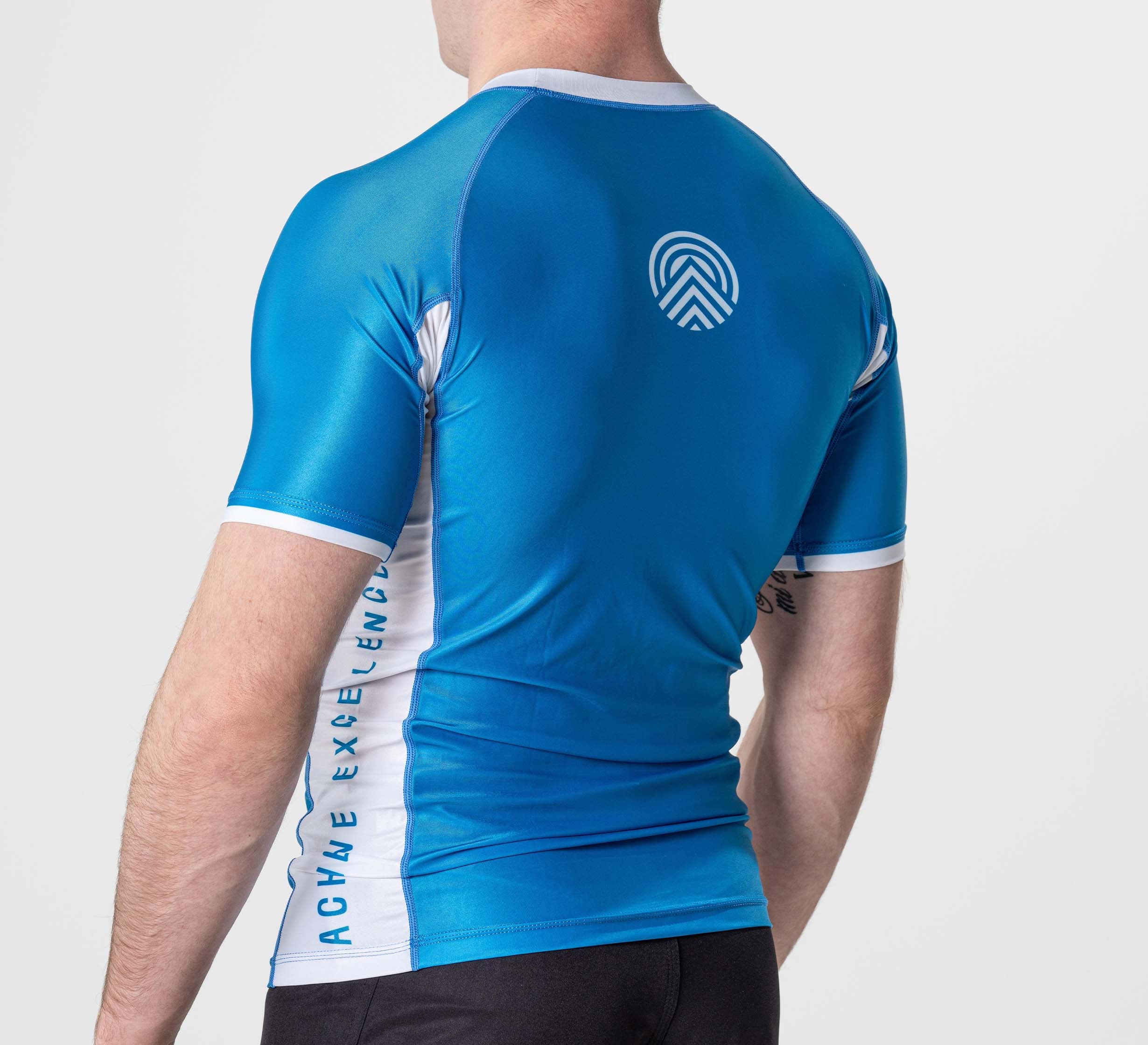 A person is seen from the back wearing the BJJ Flex Lite Rashguard Blue, a blue and white, short-sleeve, athletic, tight-fitting shirt with a circular logo on the upper back and "ACHVE EXCELLENCE" printed along the side panel.
