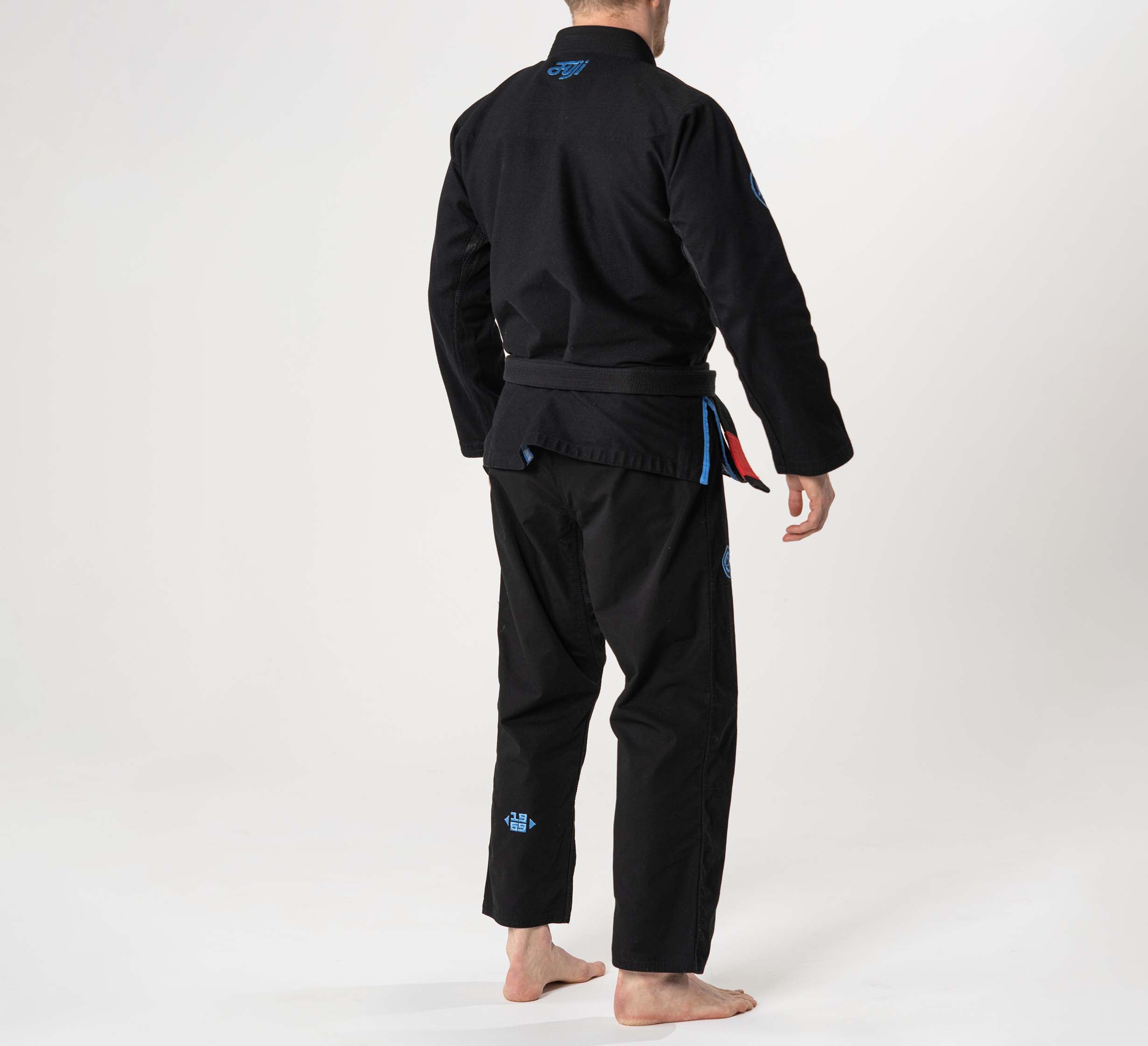 A person wears a Flow-Tech BJJ Gi in black and stands barefoot on a light background, facing away from the camera with their right arm slightly extended.