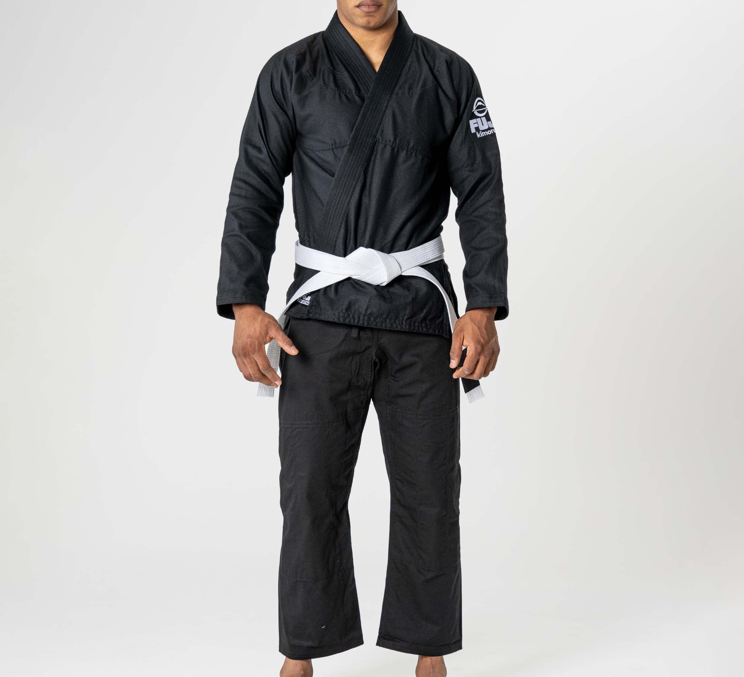 A person stands barefoot, facing forward on a white background, wearing the Starter Adult BJJ Gi Black with a white belt tied at the waist; their face is not fully visible.