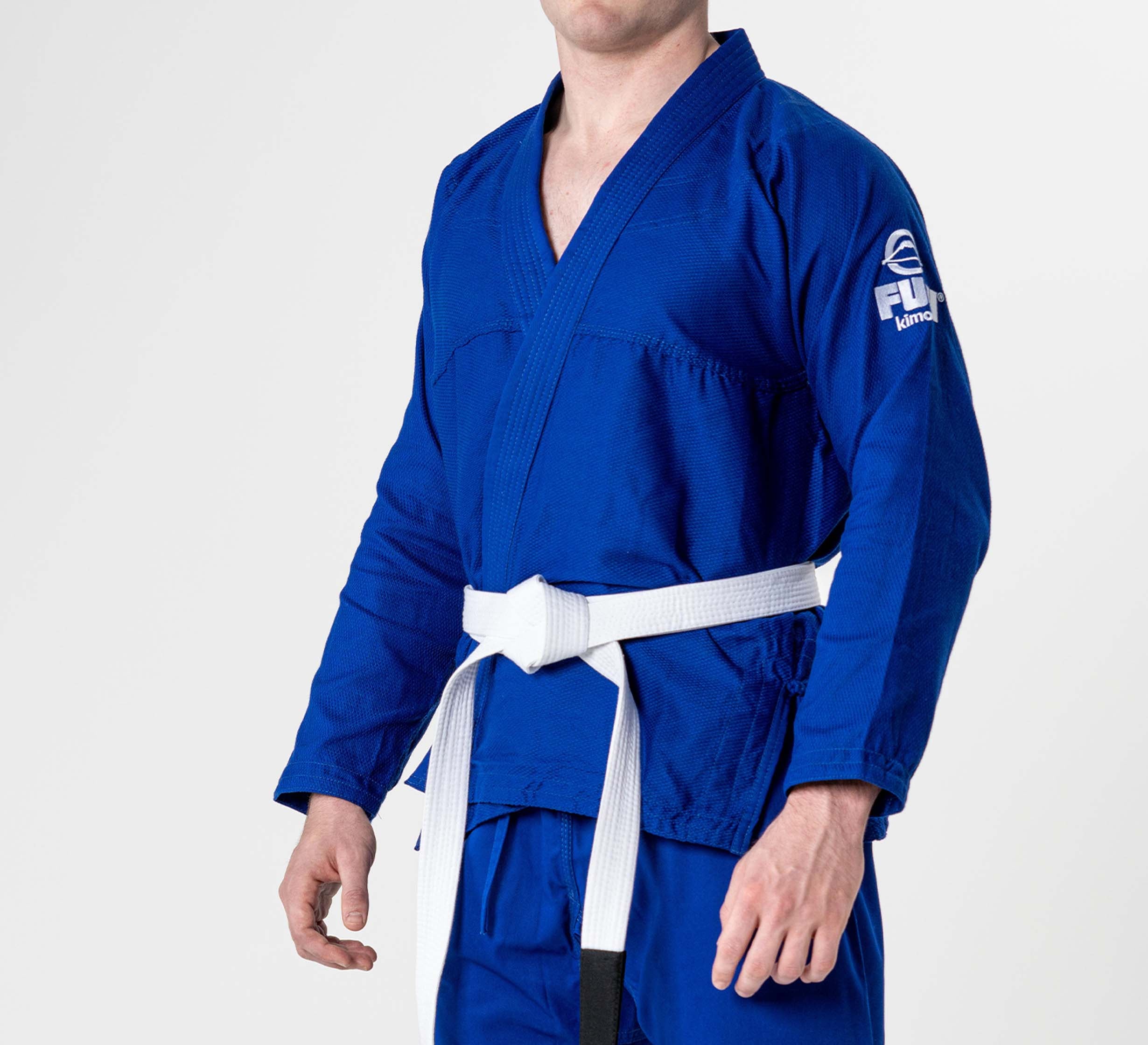 A person wearing the Starter Adult BJJ Gi Blue with a white belt, standing against a plain white background. The FUJI logo is visible on the left sleeve. The person's face is not visible.