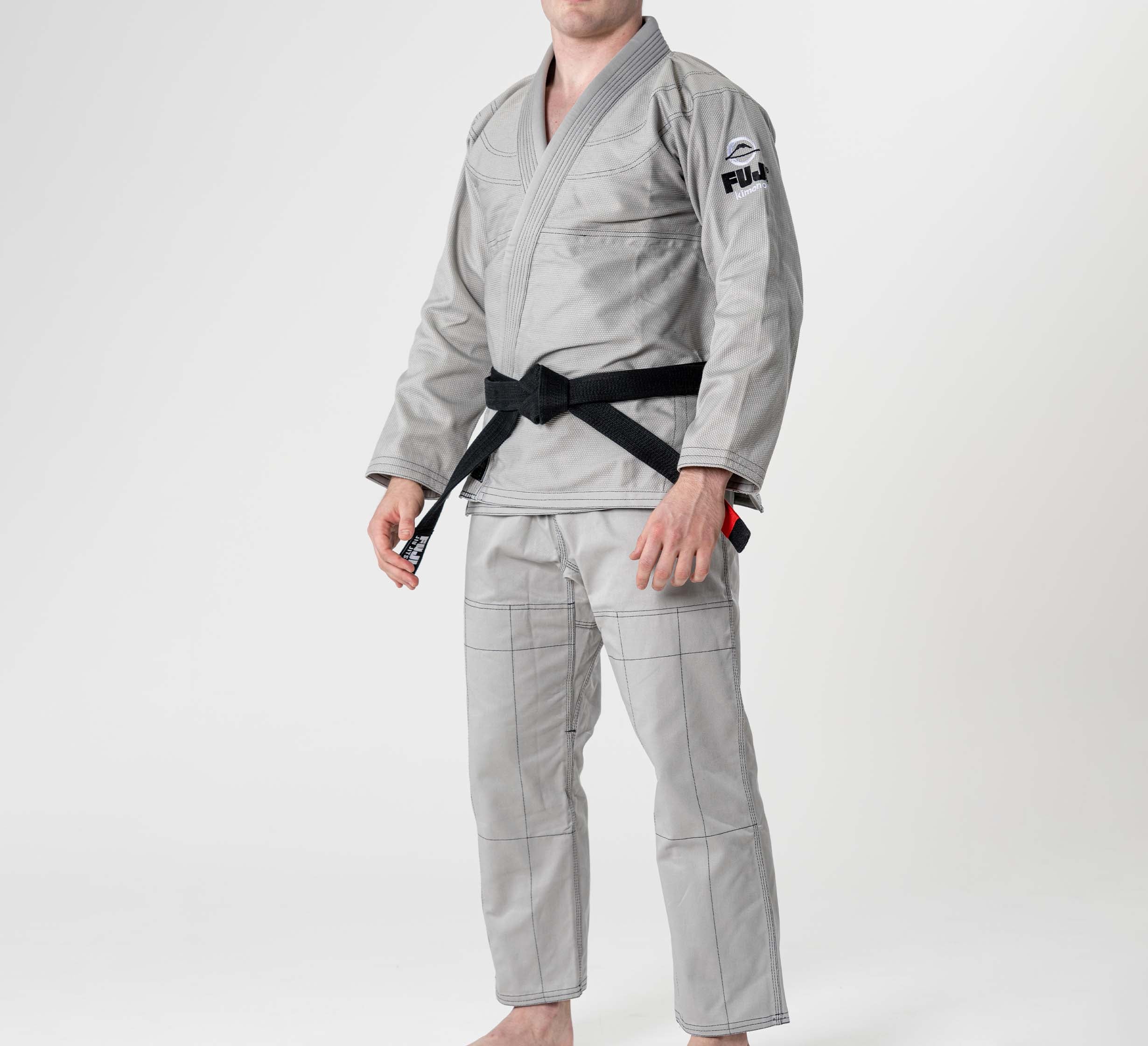 A person wearing the Lightweight BJJ Gi Grey with a black belt stands barefoot against a plain white background. The gi features visible stitching and a patch on the left sleeve.