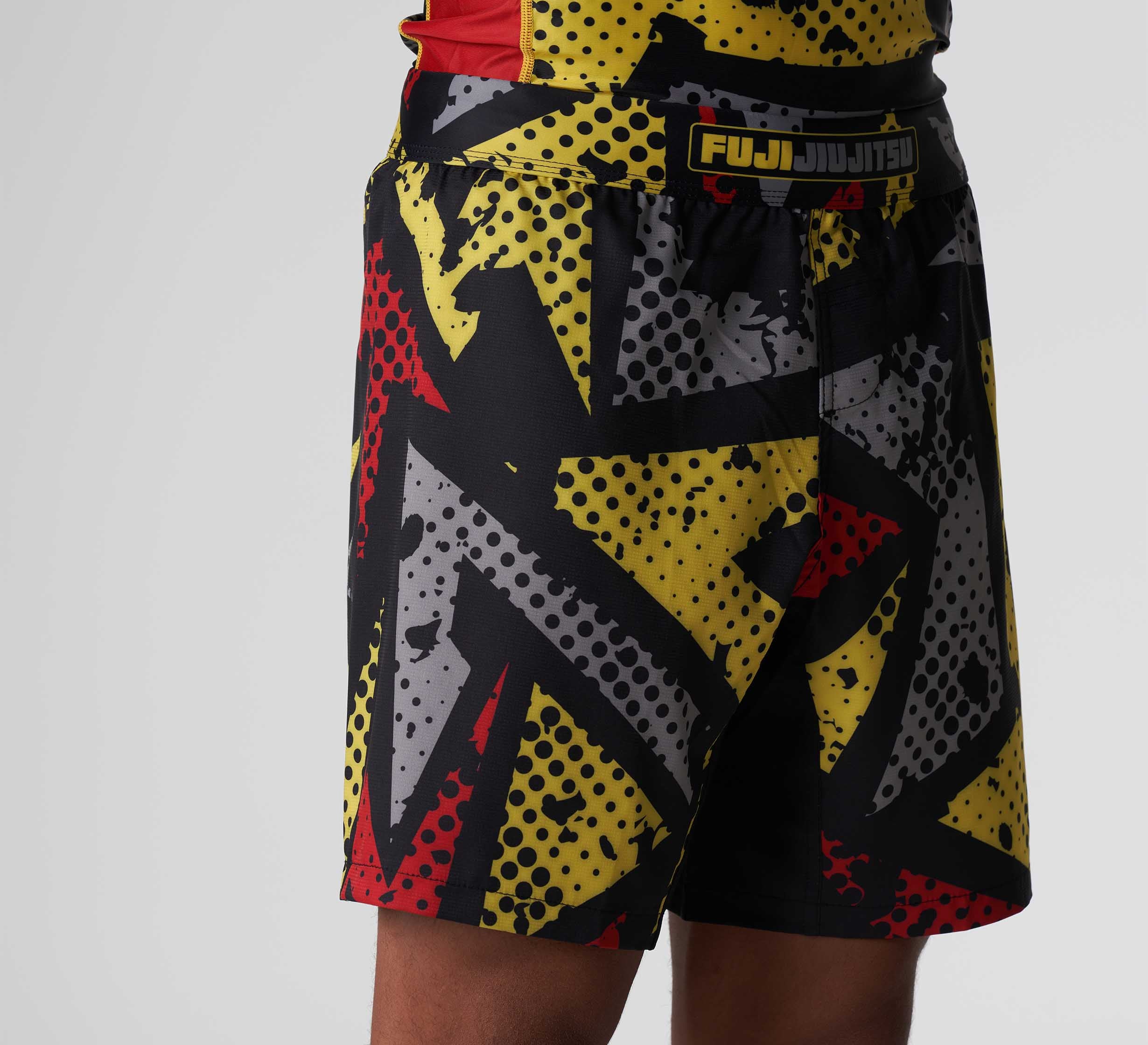 A person wearing Shuriken Flex Lite Shorts Black/Yellow with bold comic-style graphics, shown from waist to mid-thigh against a plain gray background.