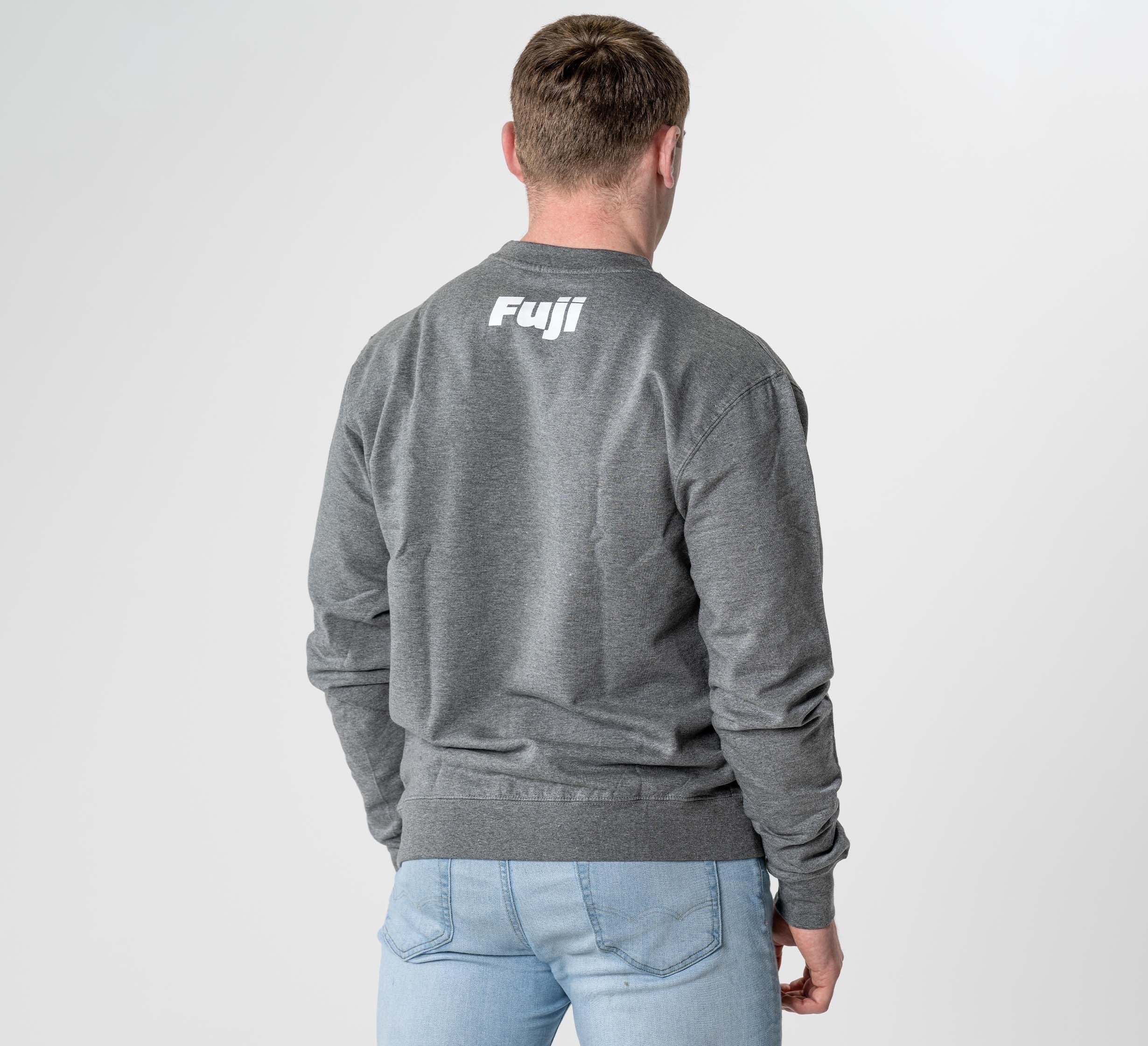 A man with light brown hair wears the Jiu Jitsu Player Crewneck Heather Grey, featuring "Fuji" in white letters on the back, paired with light blue jeans. He stands facing away from the camera against a plain white background.