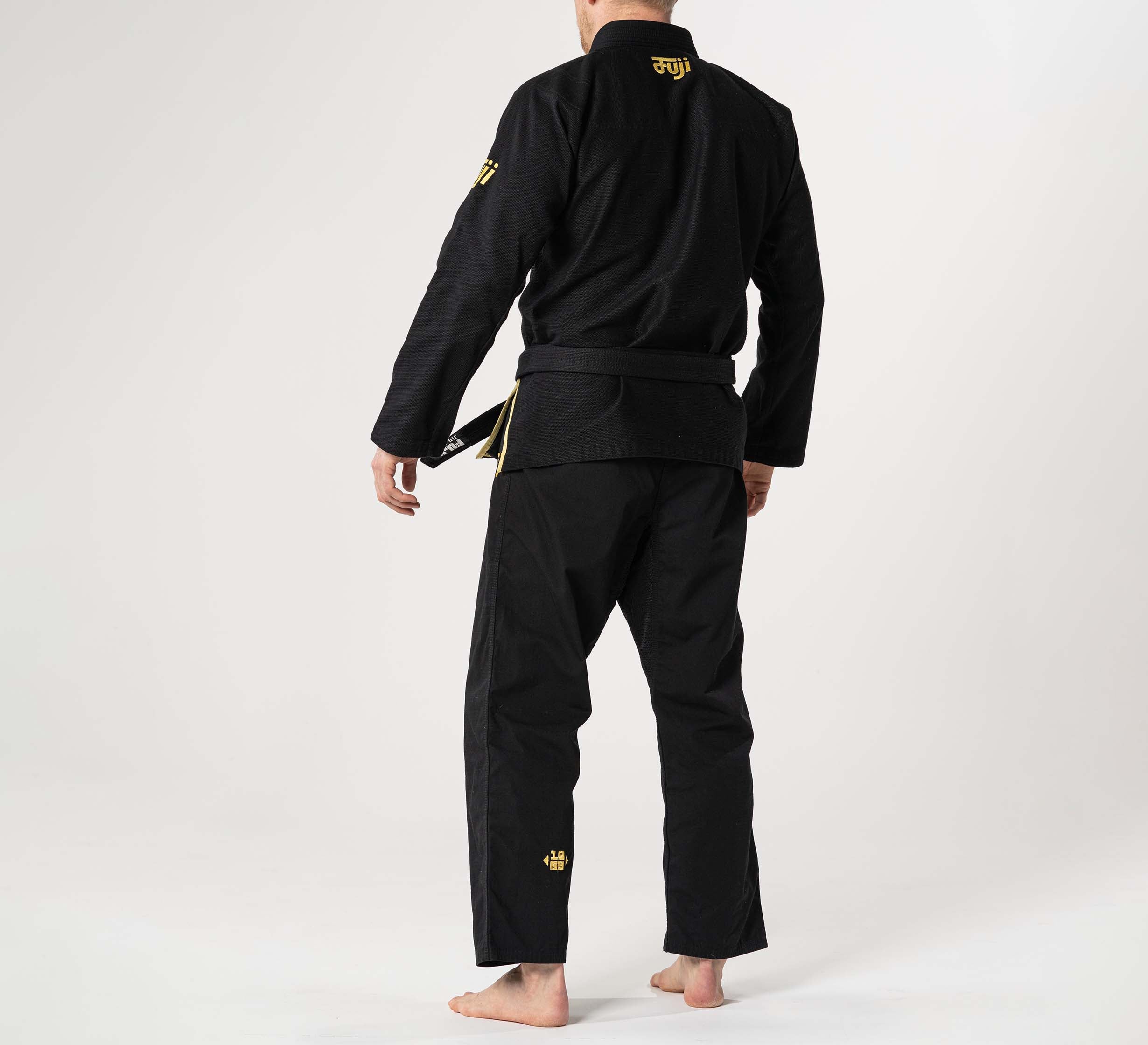 A person stands barefoot on a white surface, wearing the Flow-Tech BJJ Gi Black/Gold with gold lettering and small gold logos, facing away to display the back of the uniform.