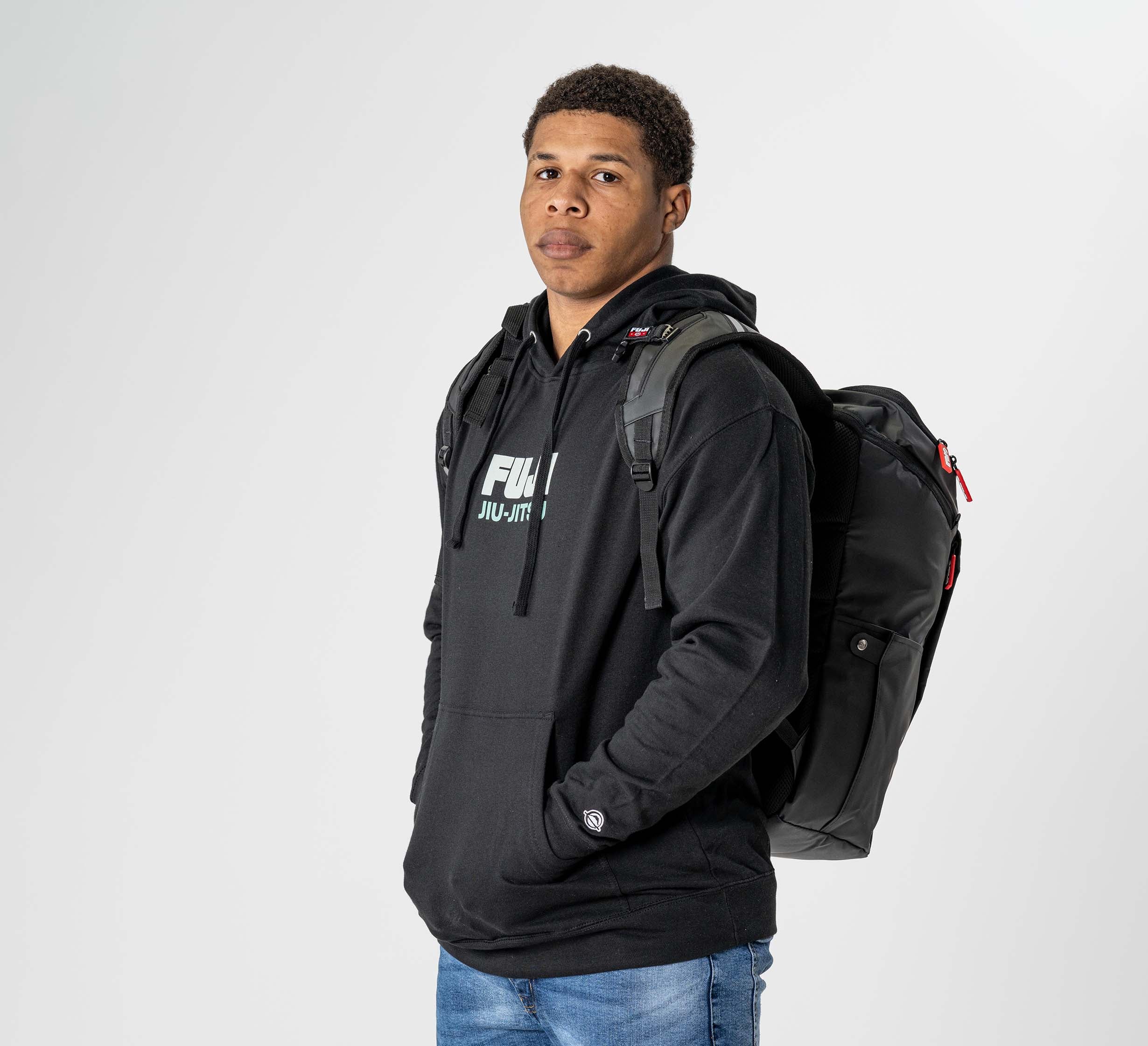 A person wearing a black hoodie and blue jeans stands sideways, modeling the Urban Day Backpack Black, looking at the camera against a plain white background.