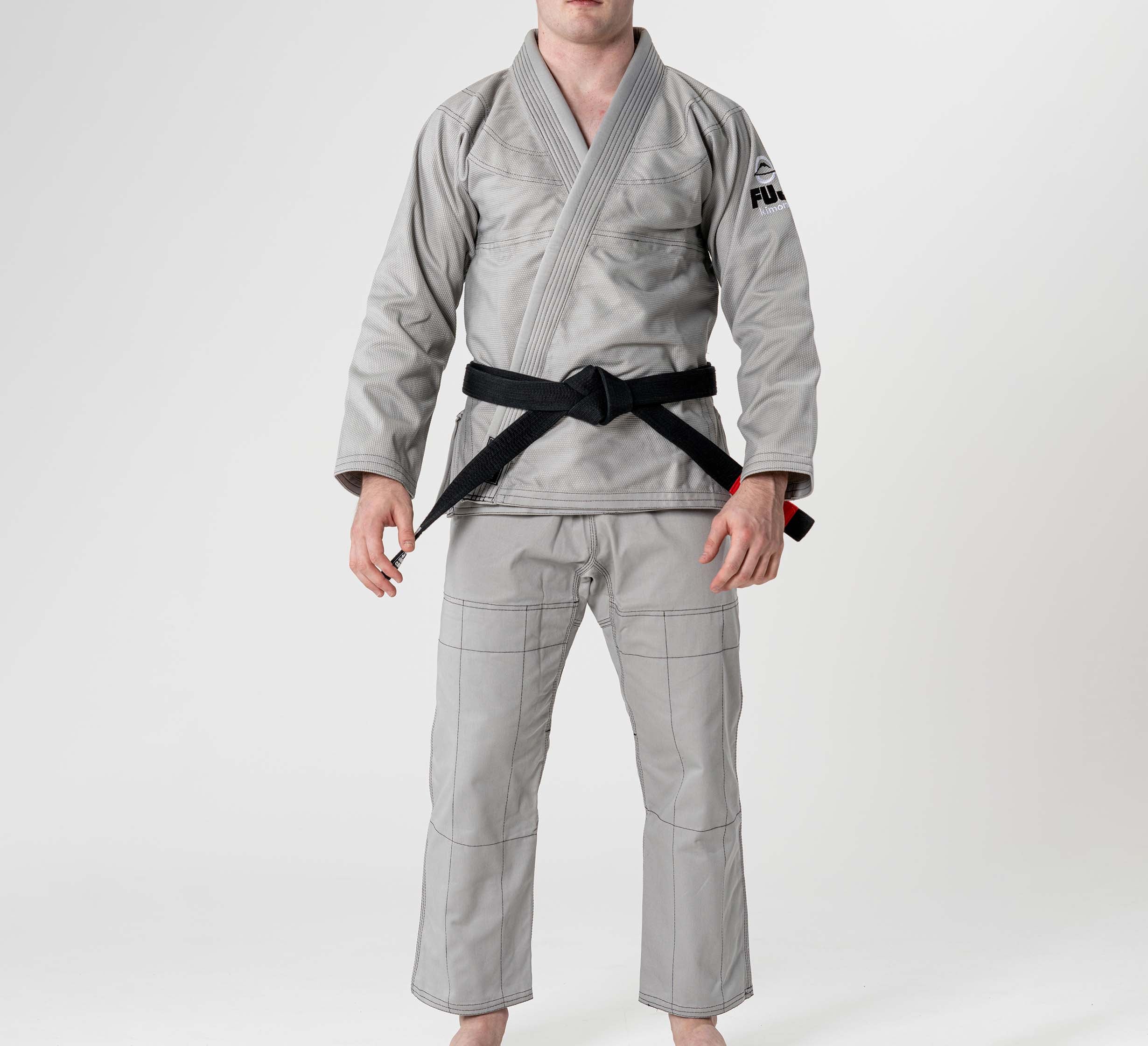 A person stands barefoot wearing the Lightweight BJJ Gi Grey with a black belt, against a plain white background. The head is not visible in the image.