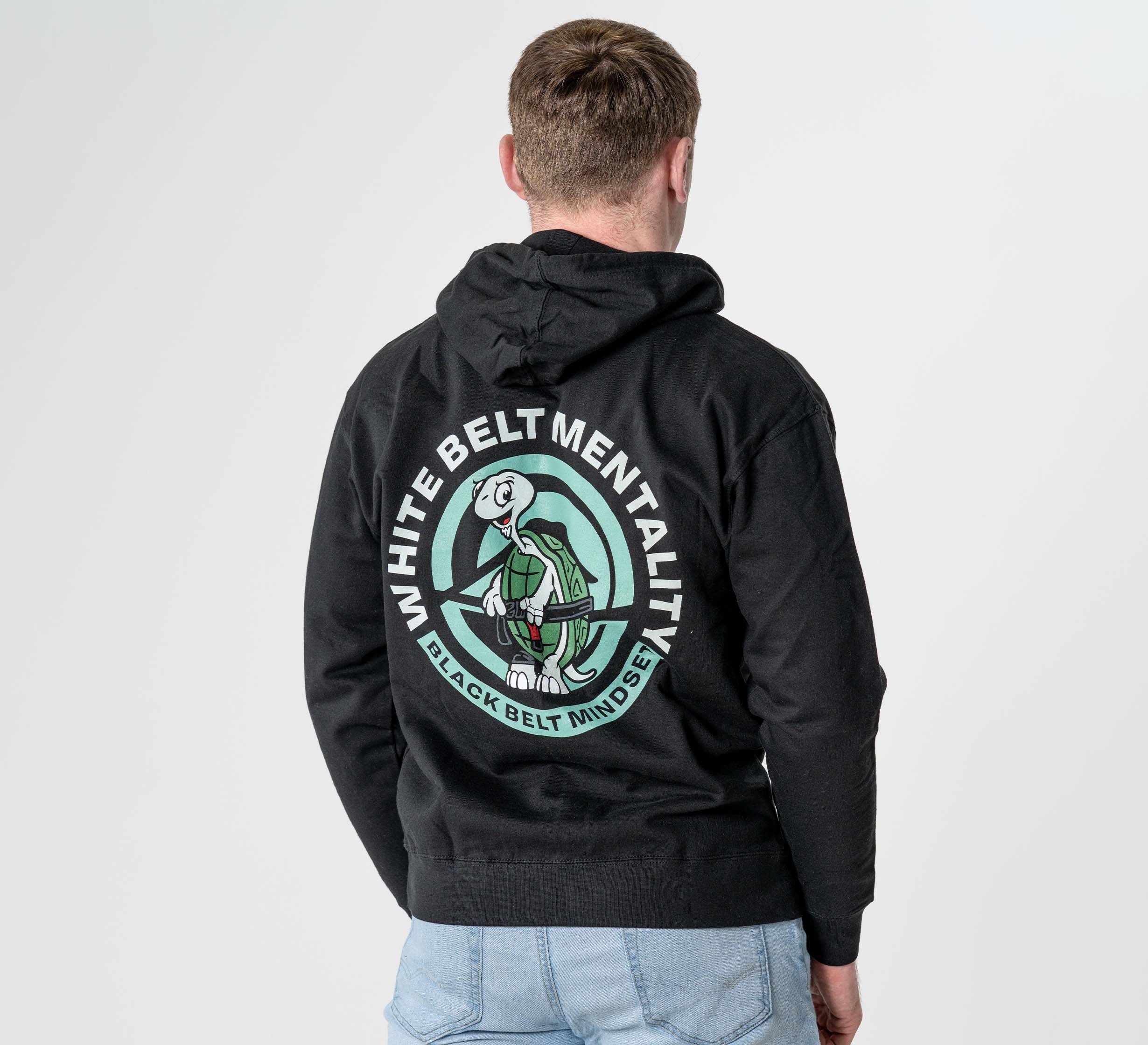 A man faces away, wearing the Black Belt Mentality Hoodie Black with a cartoon martial artist graphic and “WHITE BELT MENTALITY, BLACK BELT MINDSET” text on the back. He has short hair and light blue jeans.