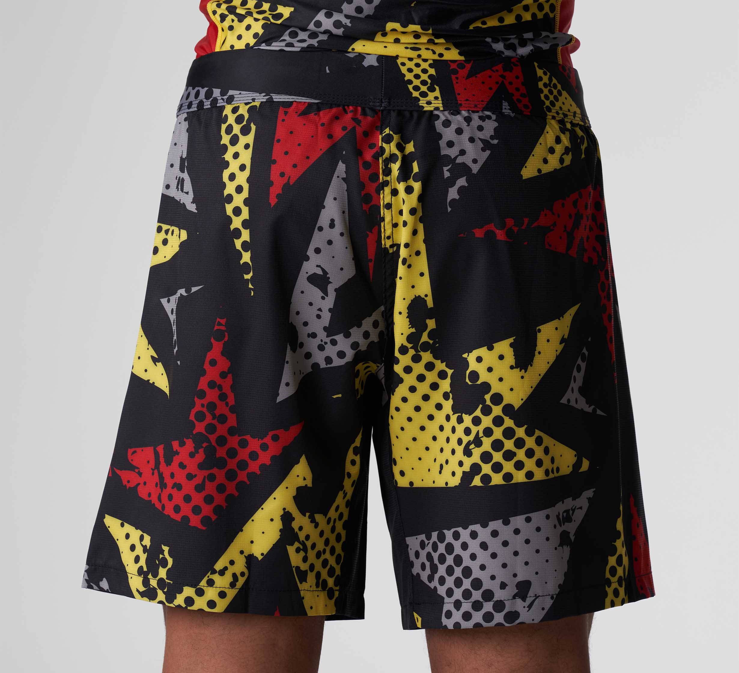 A person shown from the waist down, facing away, wears Shuriken Flex Lite Shorts in Black/Yellow featuring a bold comic-style print with red, yellow, white, and gray dots and shapes.