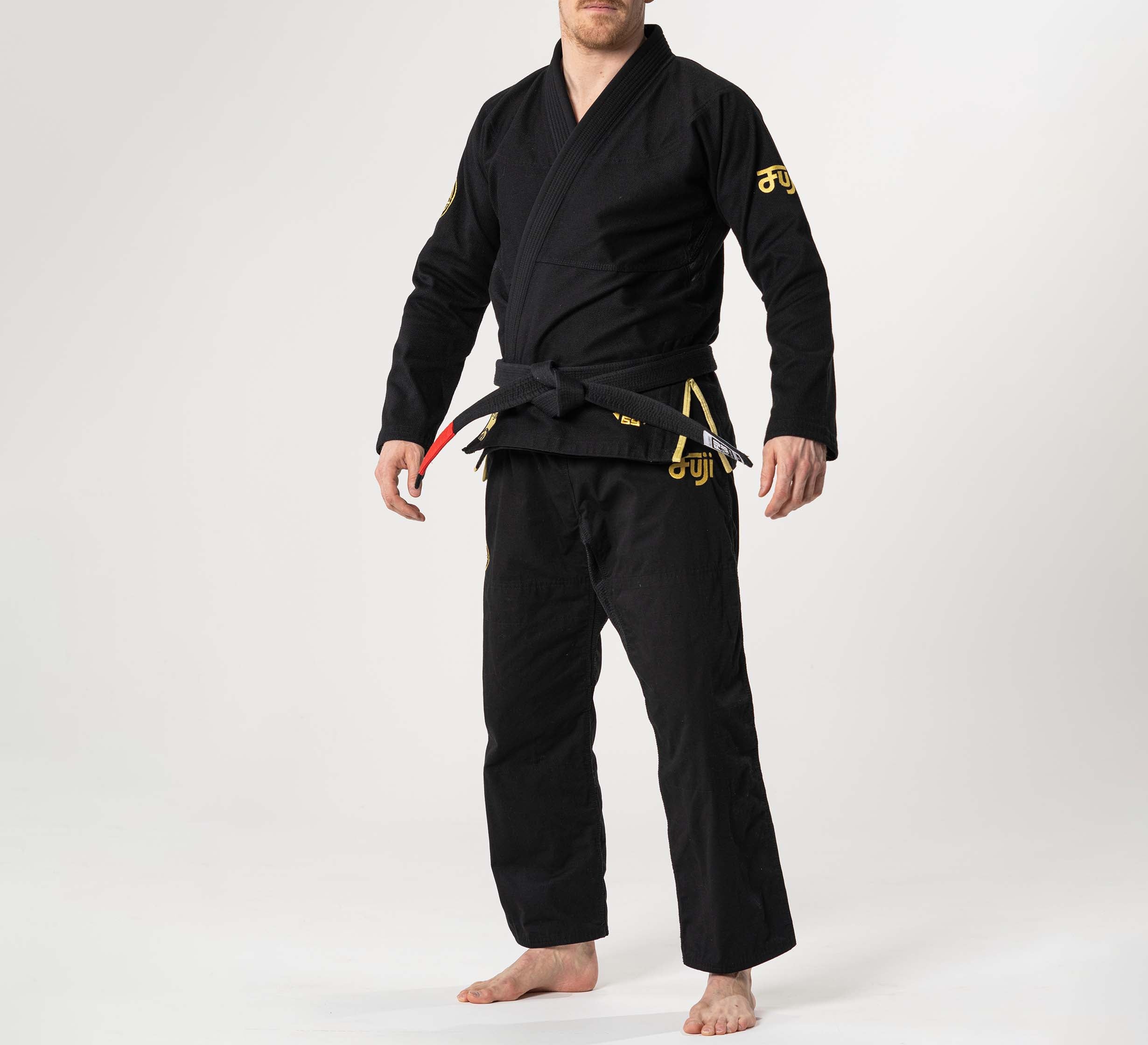 A person stands barefoot on a white background, wearing the Flow-Tech BJJ Gi Black/Gold with yellow embroidery and a black belt tied at the waist. The person's face is partially out of frame.