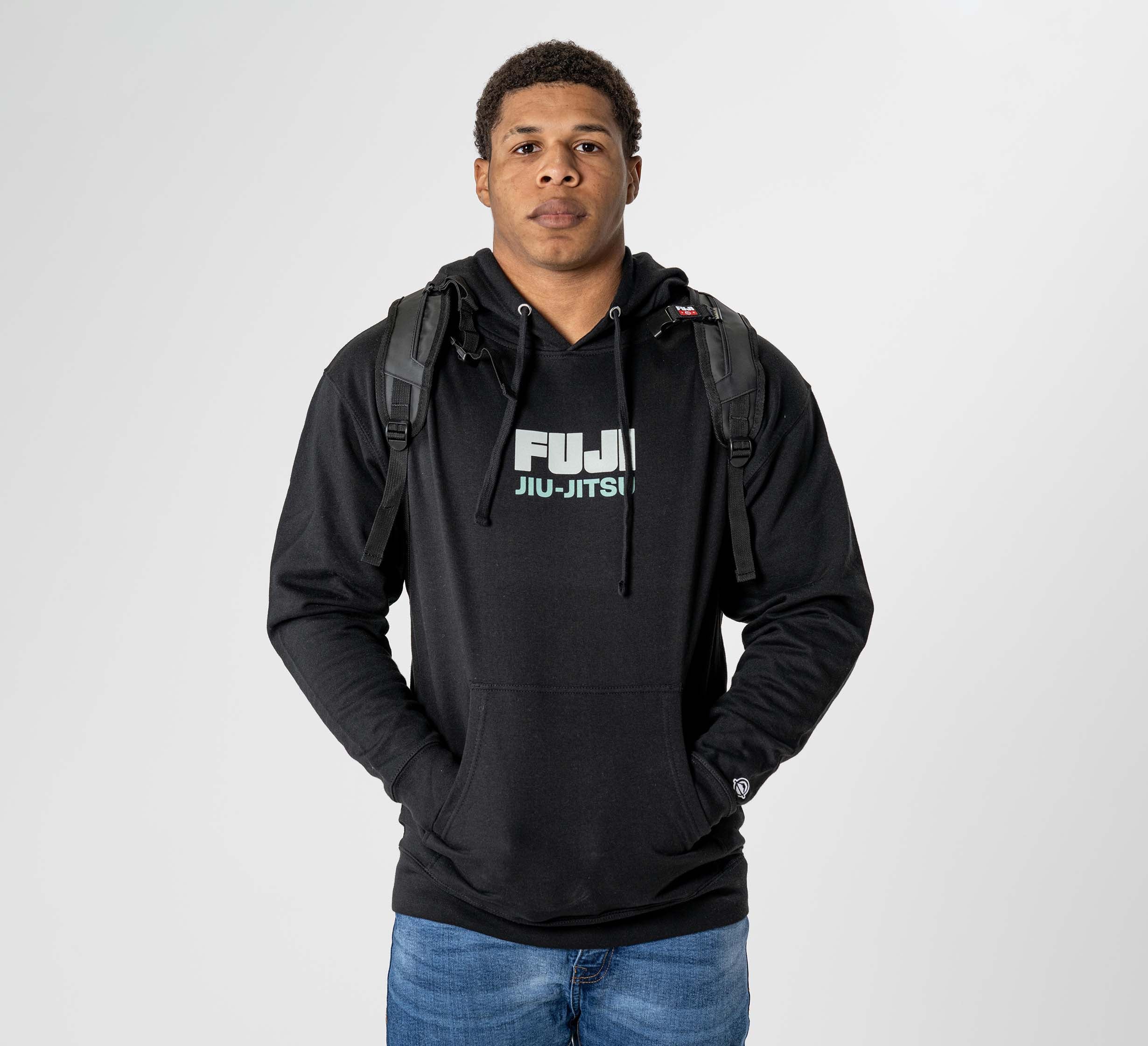 A person stands facing forward with hands in the front pocket of a black FUJI Jiu-Jitsu hoodie and wears an Urban Day Backpack Black, against a plain light gray background.