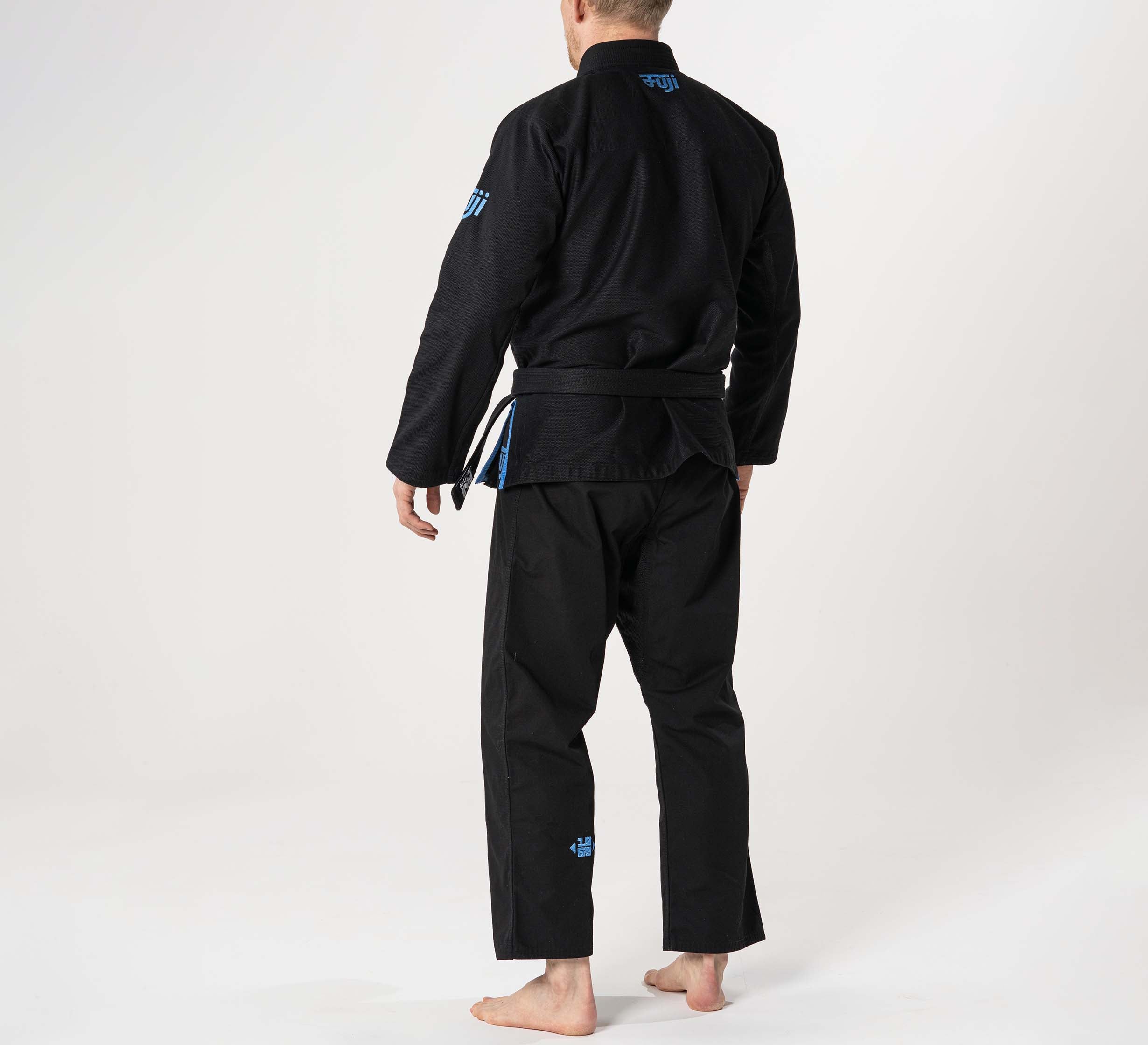 A person stands barefoot on a white background, wearing the Flow-Tech BJJ Gi Black/Blue/Grey with a belt, facing away from the camera.